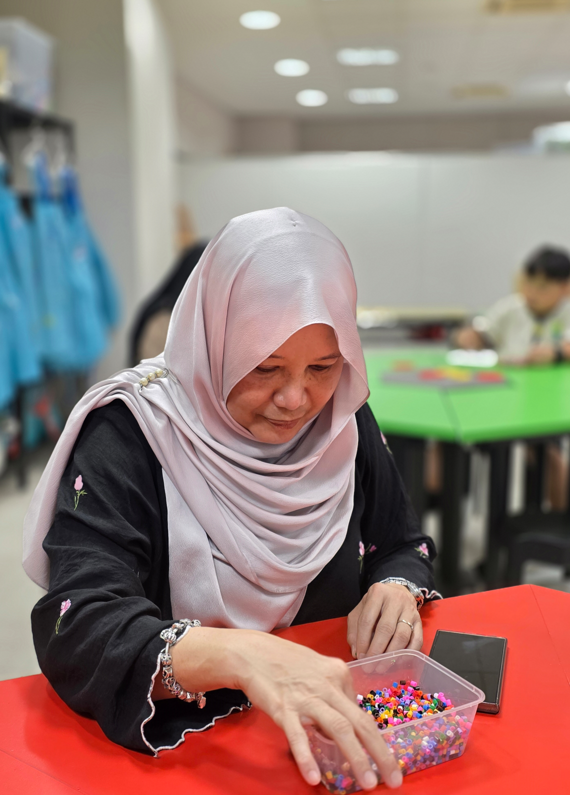 Woman wearing a pink hijab and black top sorting colorful beads in a clear container on a red table.