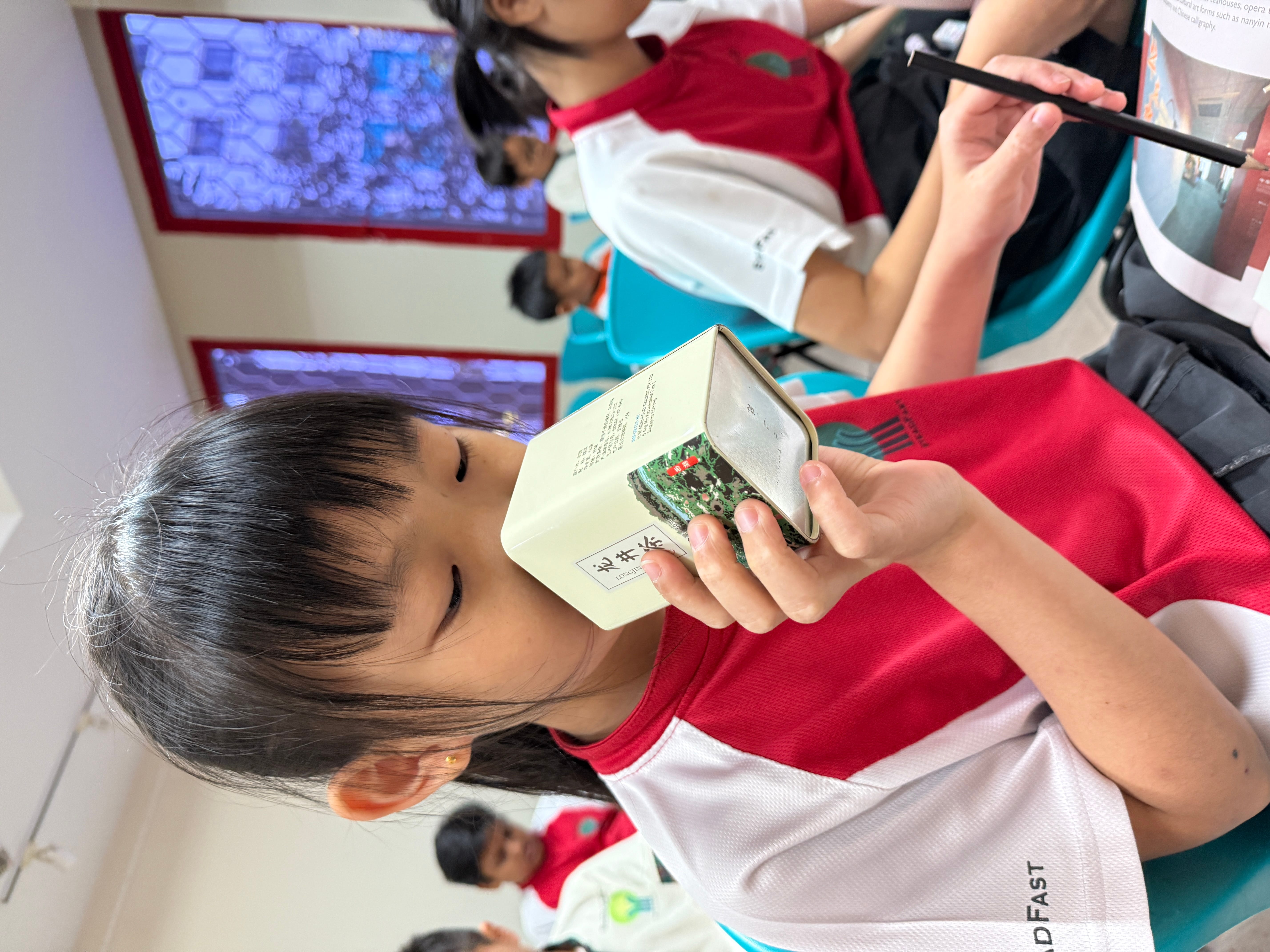 Student in red-and-white uniform holds a small circuit board box to her face in a classroom.