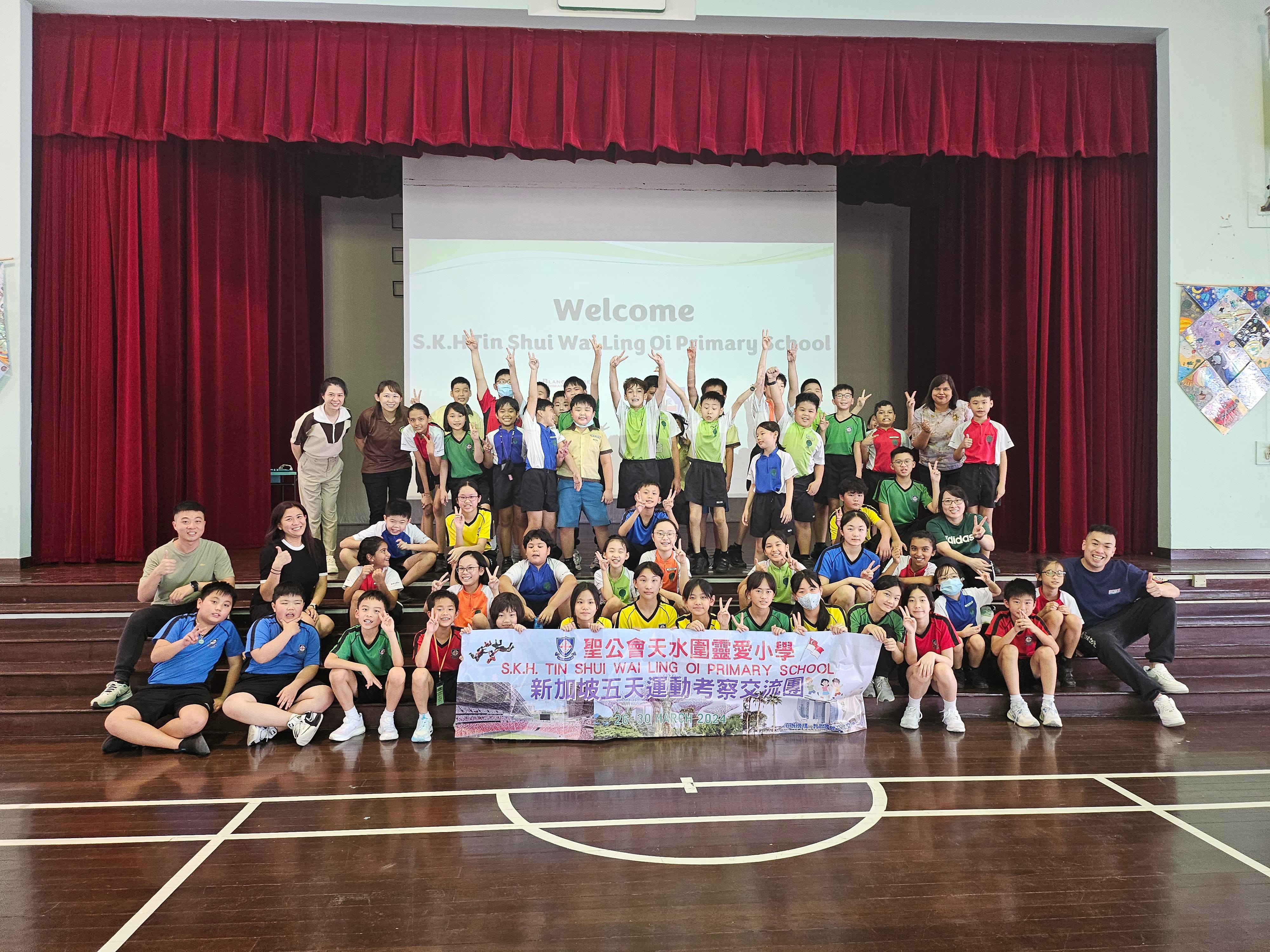 Large group of students and teachers pose on a stage with red curtains, holding a school banner.
