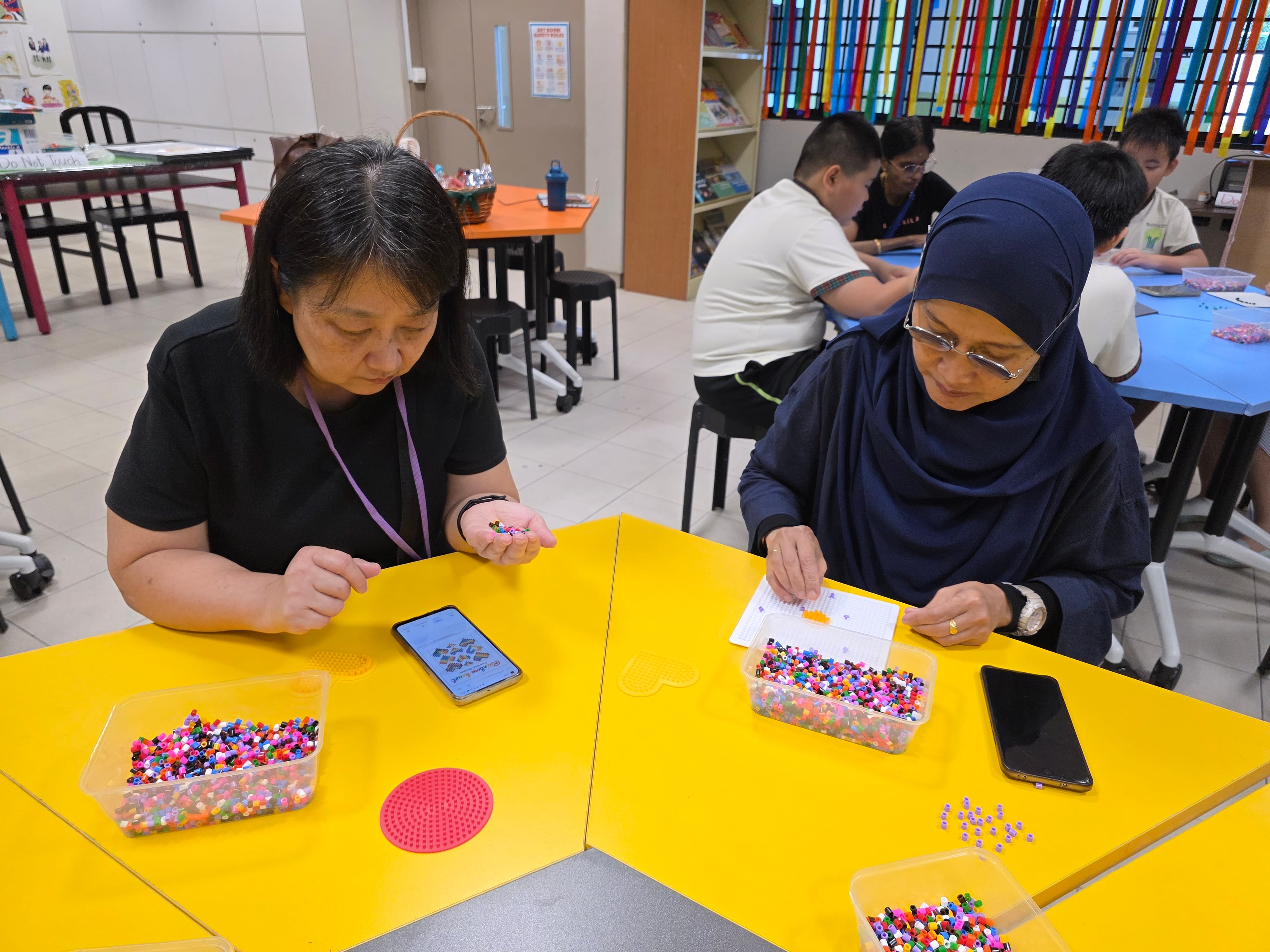 Two women sit at a yellow table sorting colorful beads, with others working in the background of a classroom.