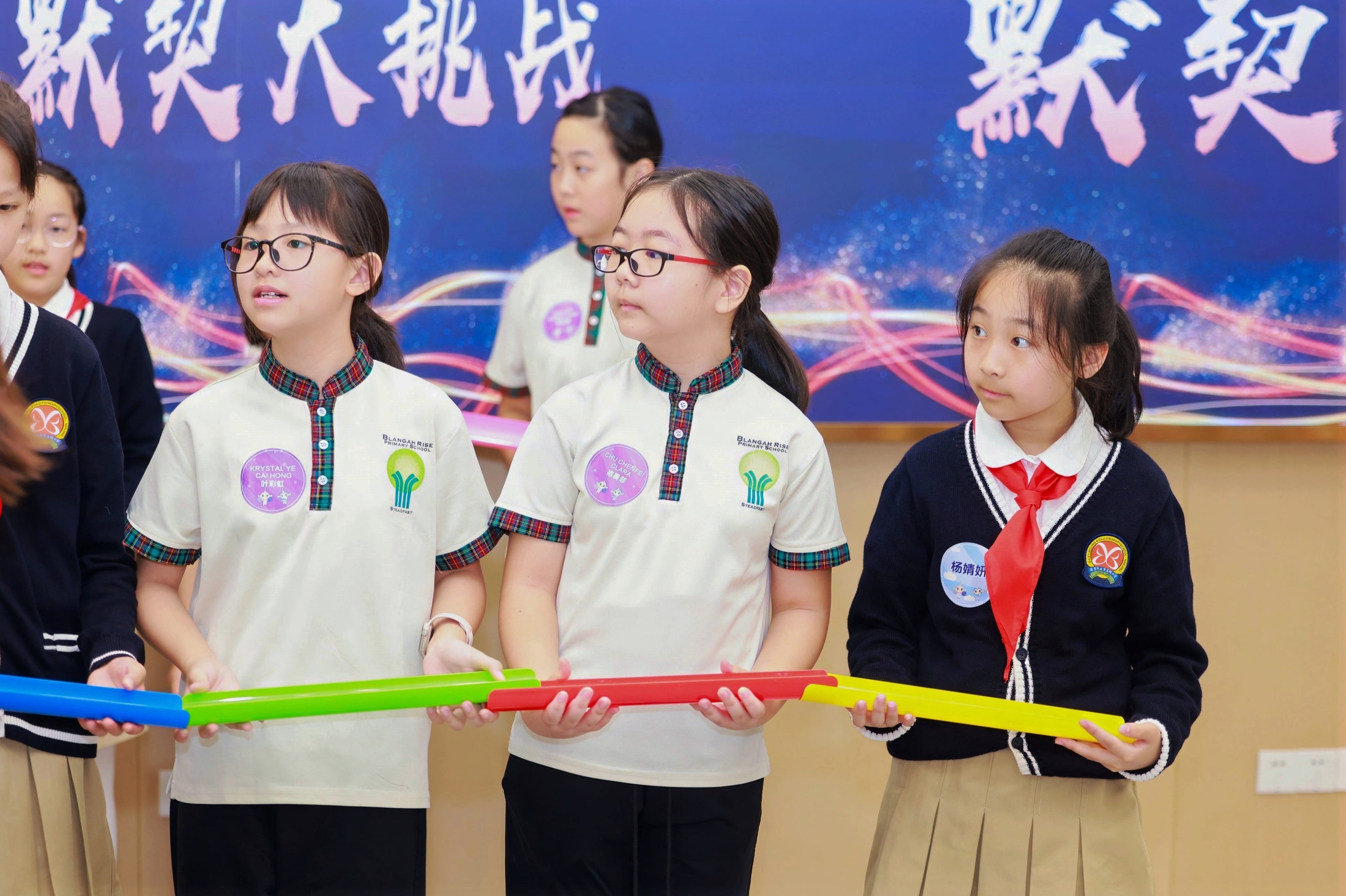 Several school children in uniforms hold colorful interlocking plastic tubes during an event with Chinese text on a banner.