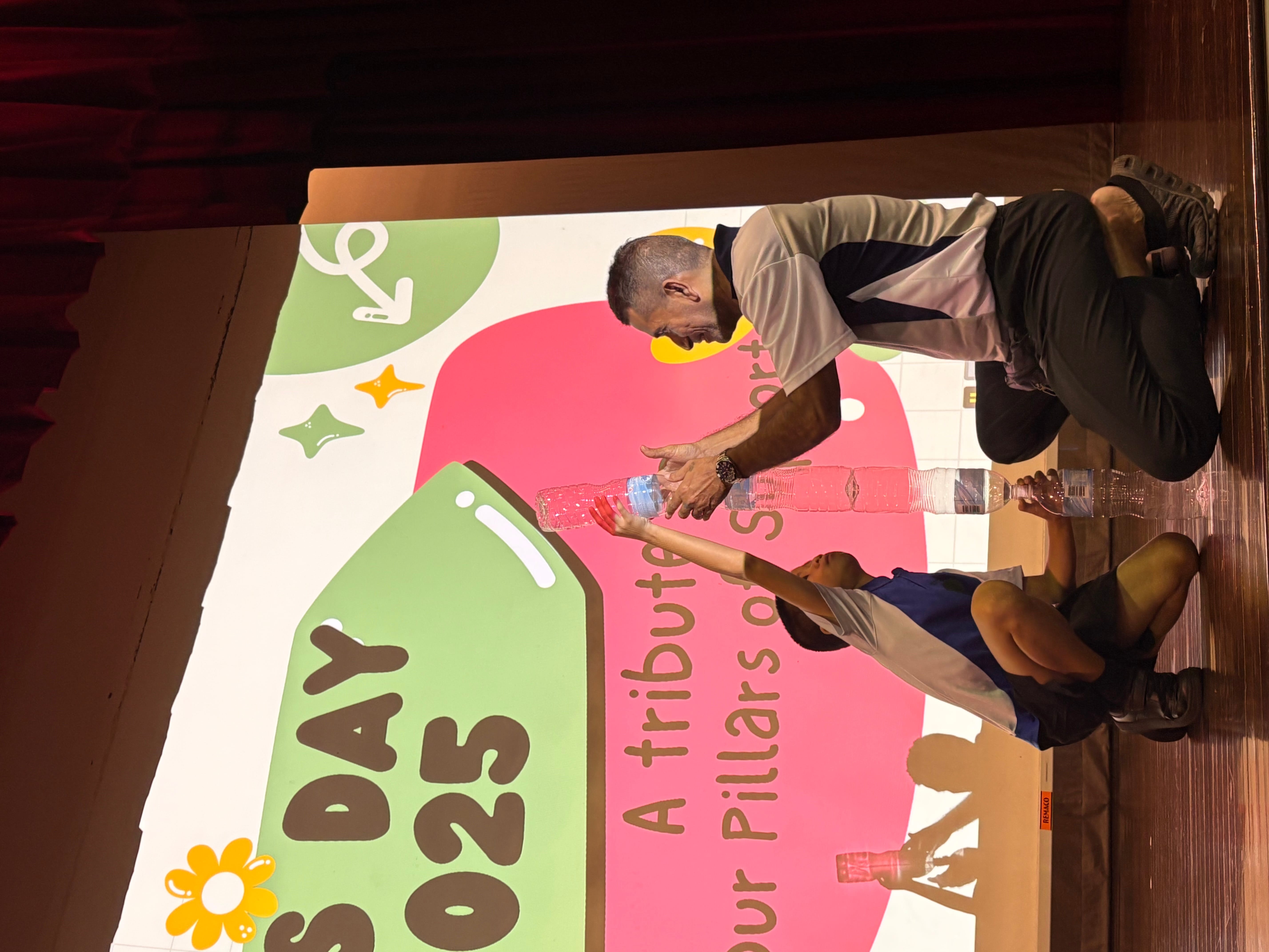 A child and an adult kneel on a stage stacking plastic bottles in front of a colorful projected slide reading "Day 2025."