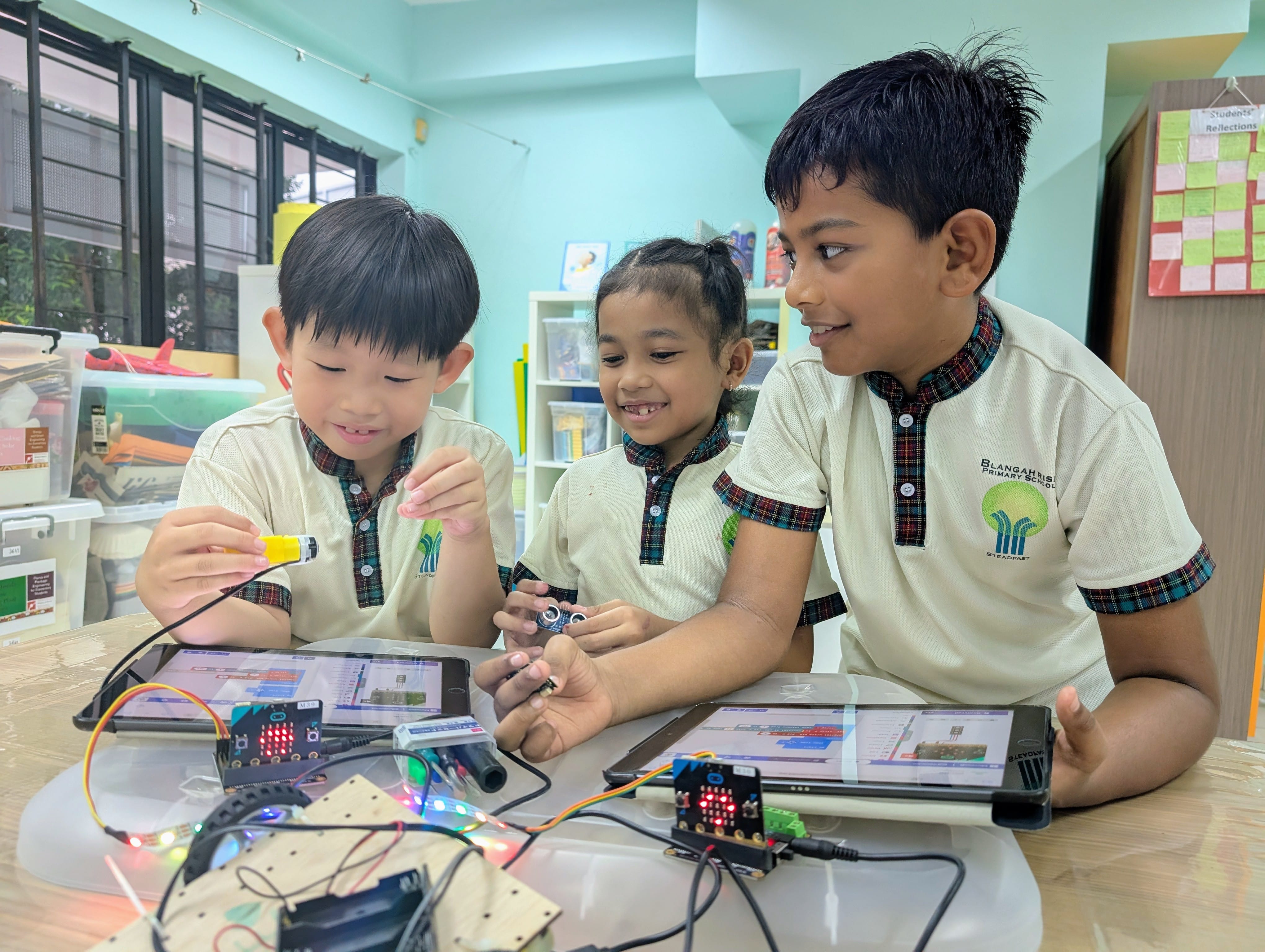 Three primary school children in uniforms work together on electronics and micro:bit circuit boards with tablets in a classroom.