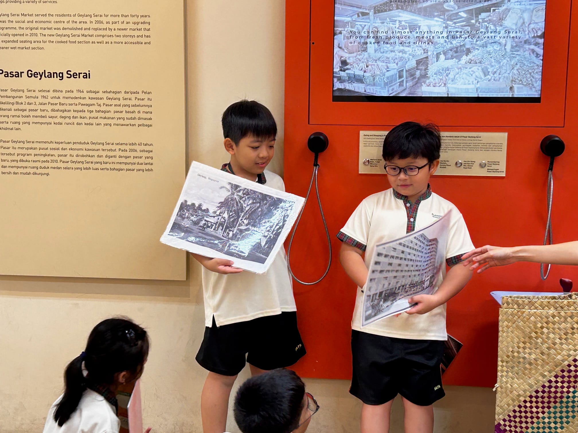 Two boys in school uniforms hold black-and-white printed photos inside a museum exhibit about Pasar Geylang Serai.