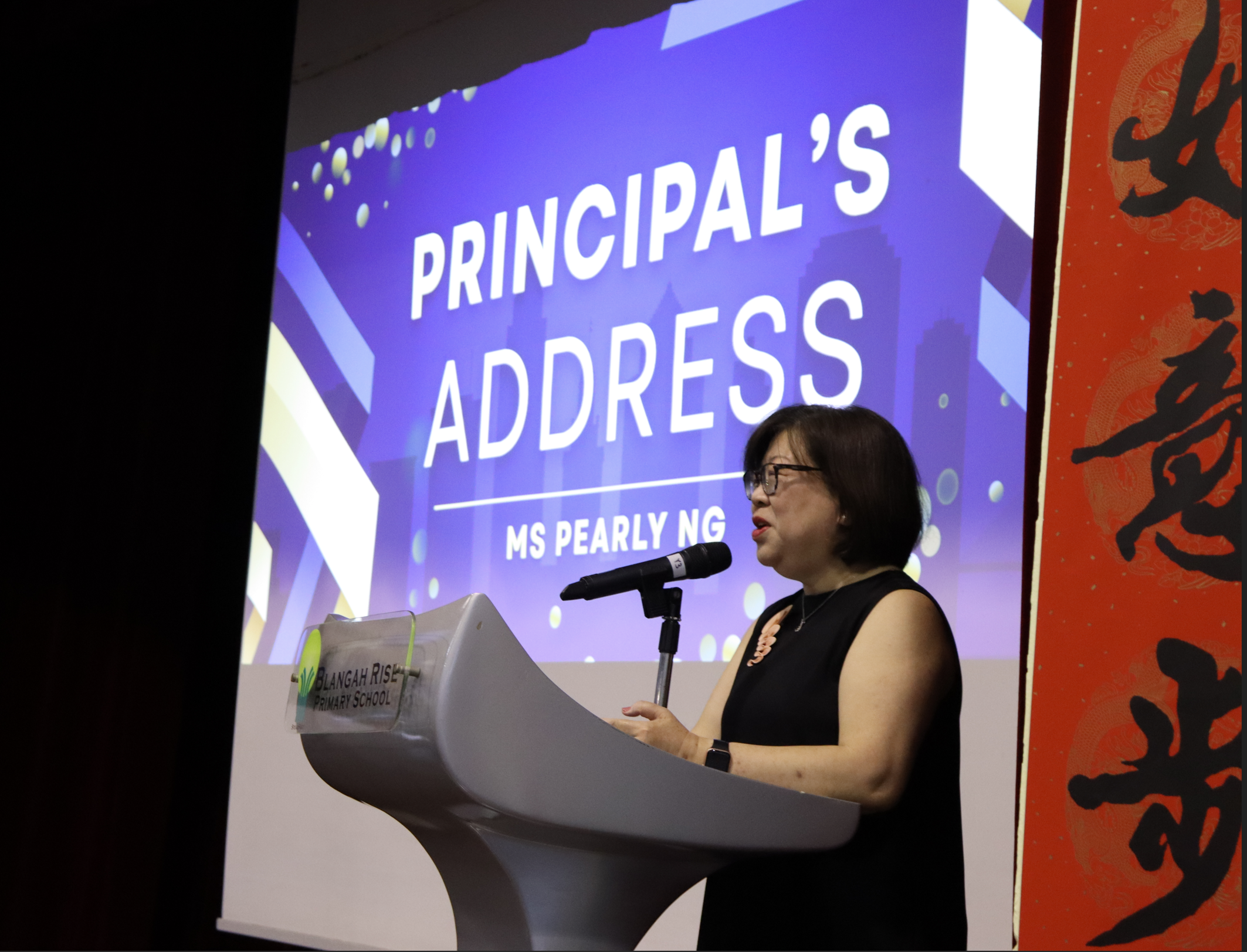 Woman speaks at a podium into a microphone, with screen behind reading PRINCIPAL'S ADDRESS MS PEARLY NG.