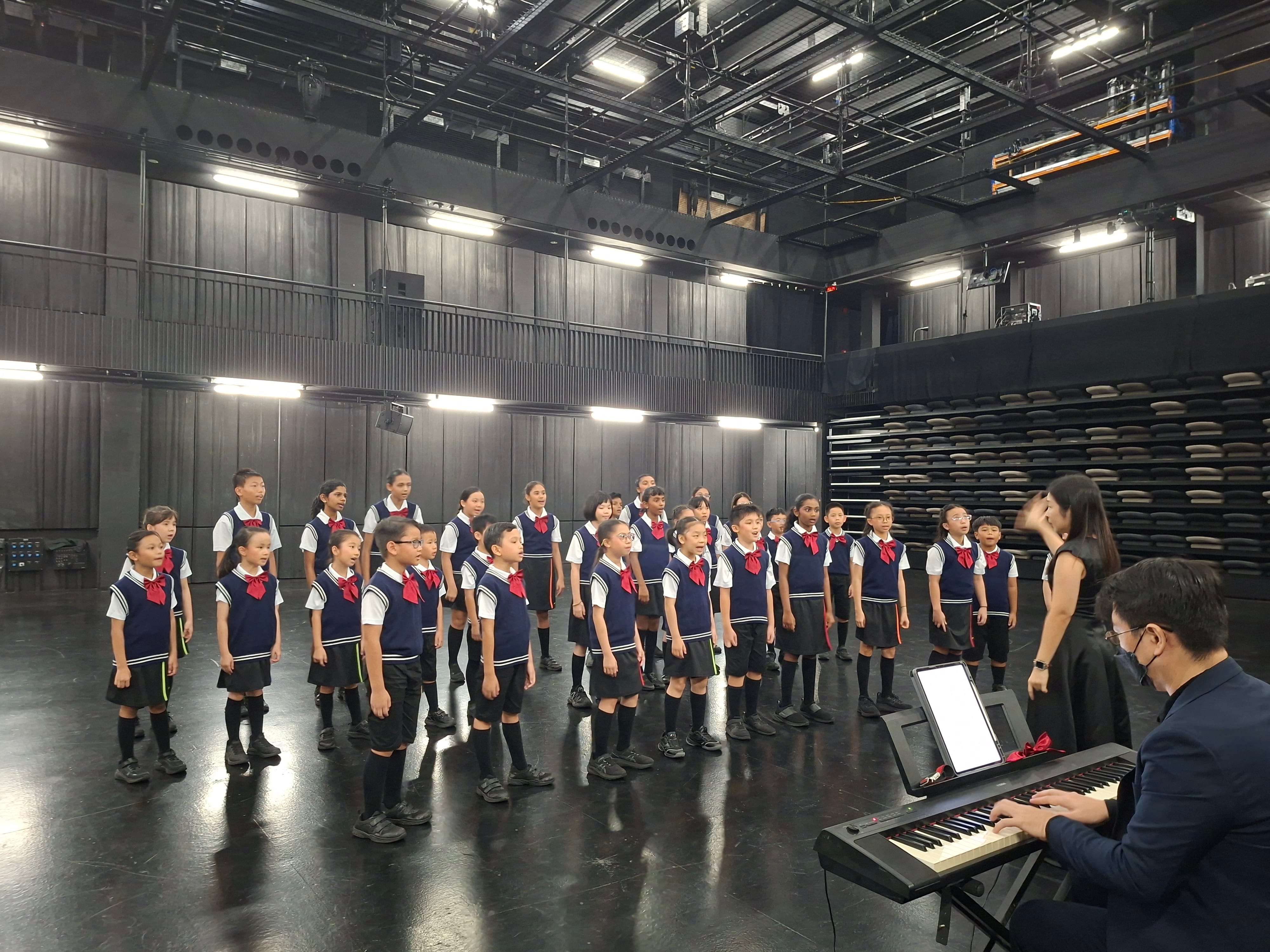 Children in navy uniforms with red ties sing in a choir on a dark stage while a conductor leads and a pianist plays.