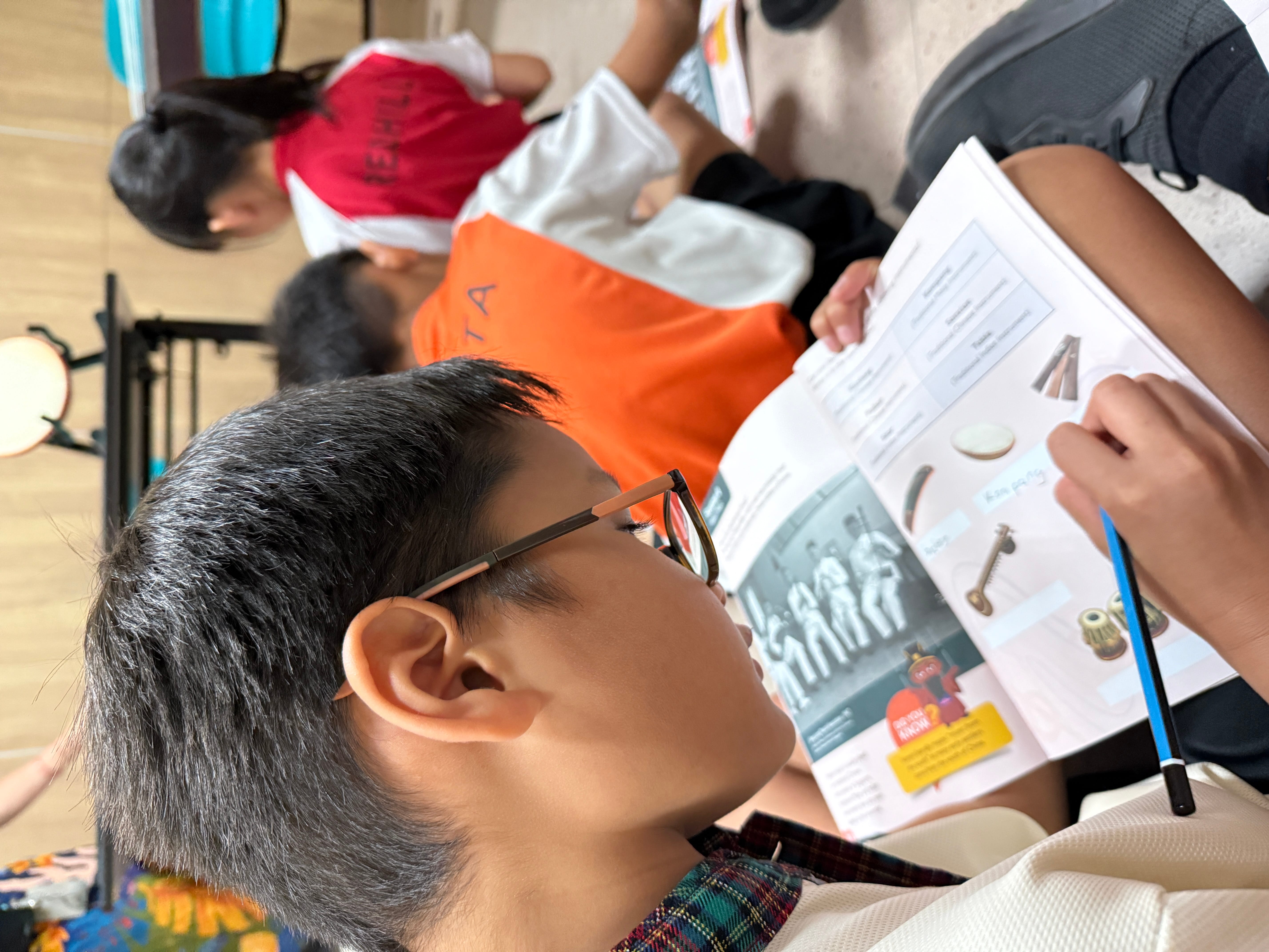 Child wearing glasses reads a workbook while other children sit nearby in a classroom.