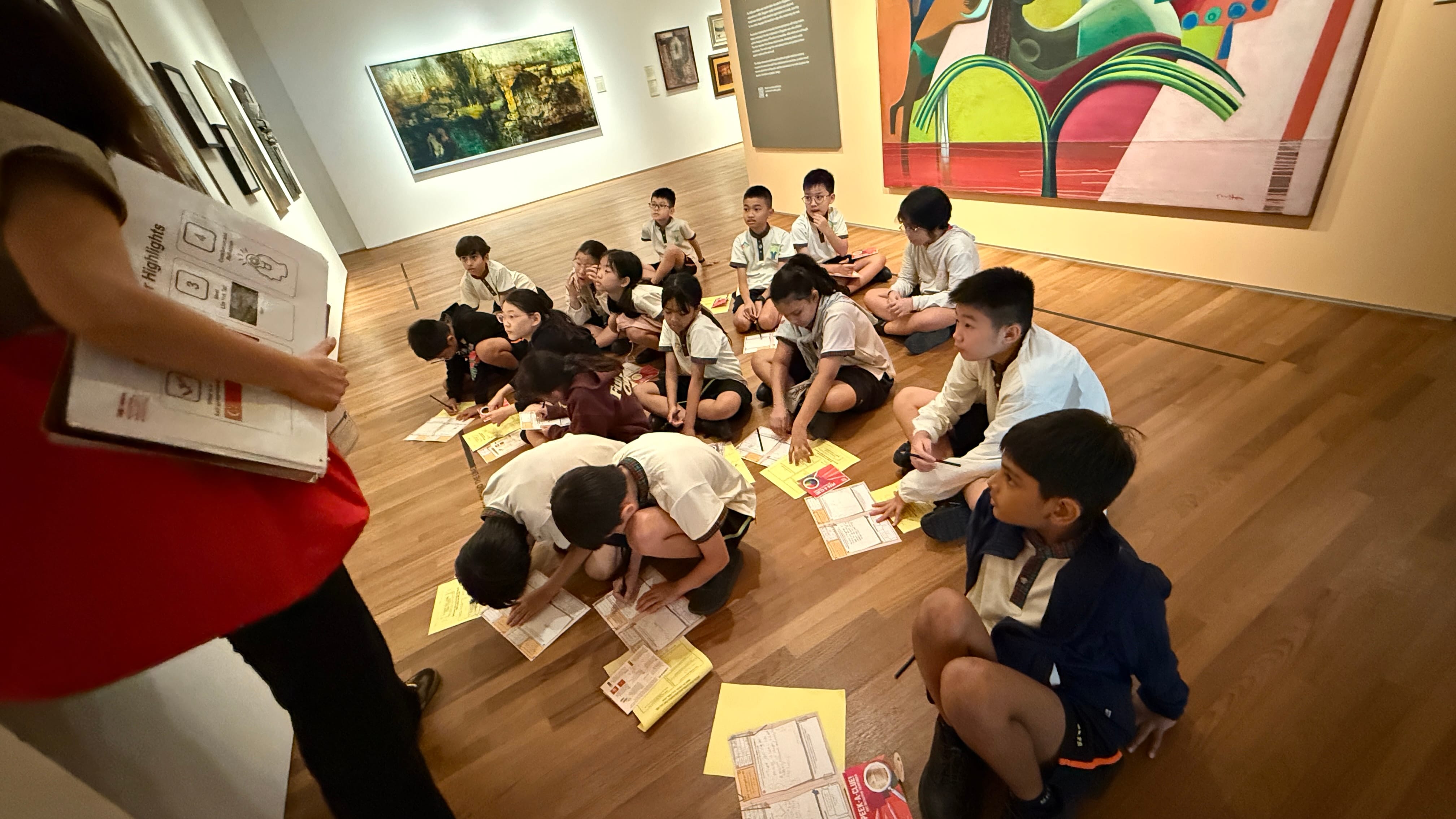 Children in school uniforms sit on a wooden gallery floor, writing on papers, surrounded by colorful artwork on walls.