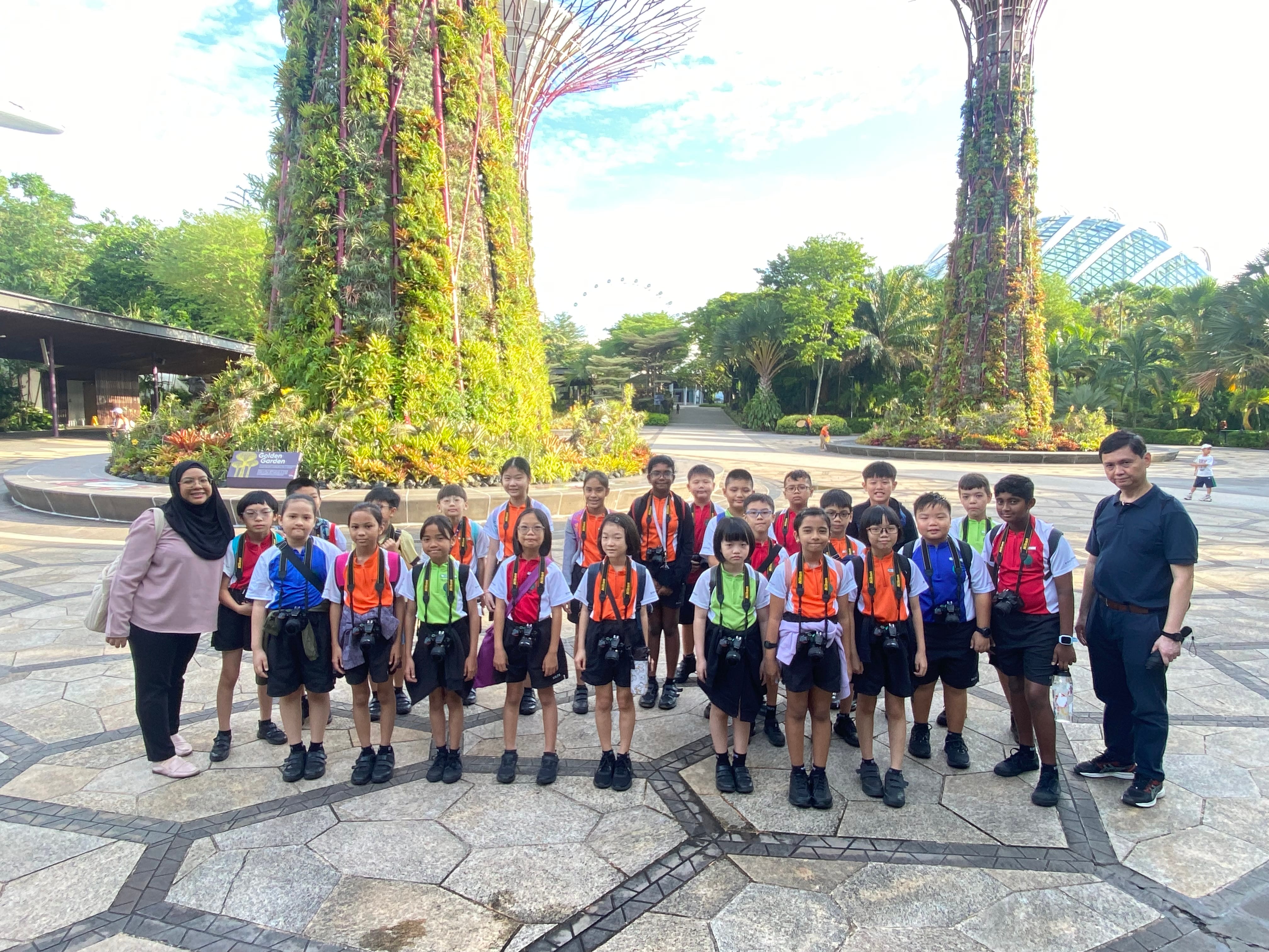 A group of school children with two adults pose in front of tall plant-covered Supertree structures at Gardens by the Bay.