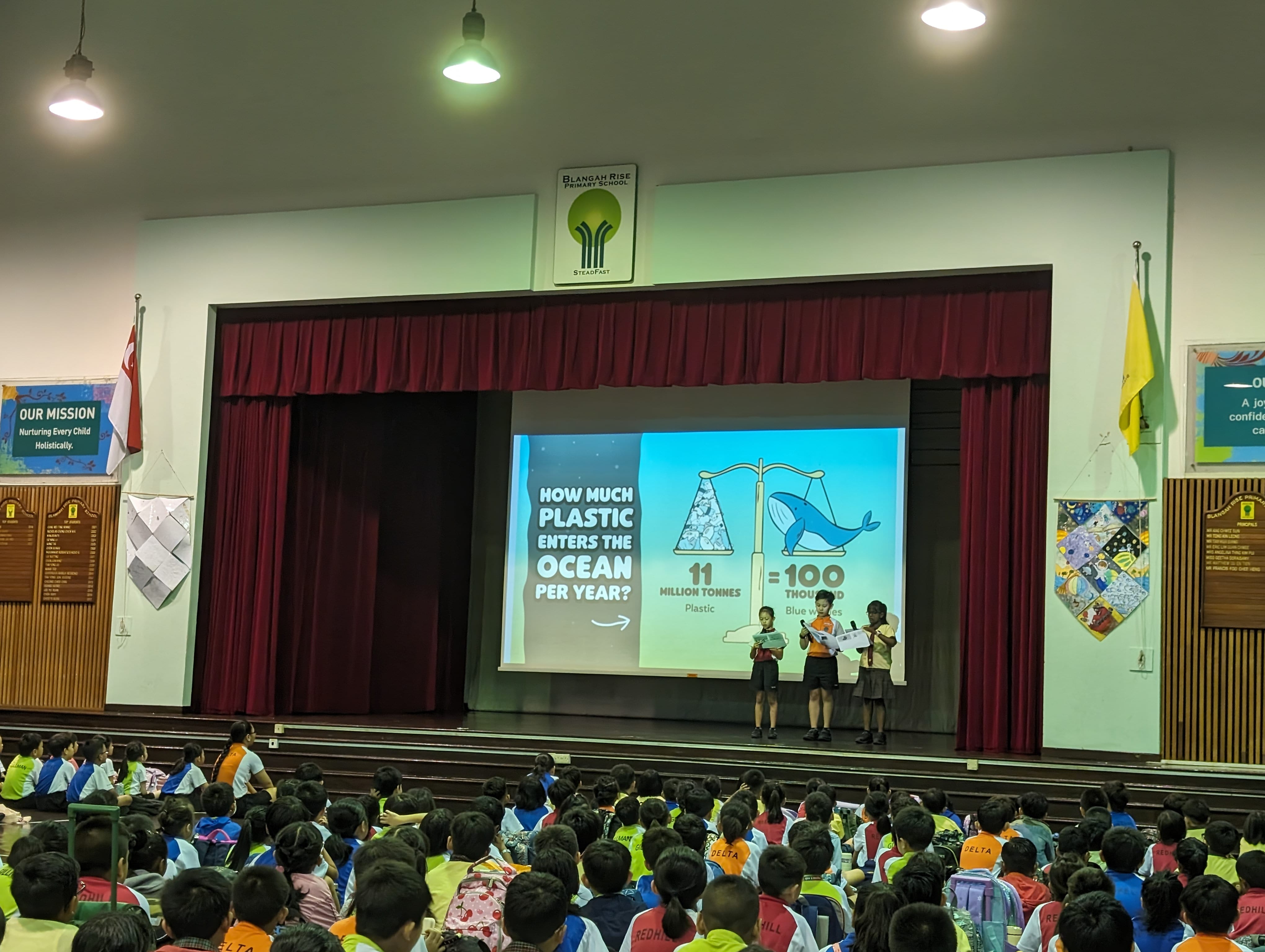 Three students present on stage at Blangah Rise Primary School with a slide about ocean plastic pollution.