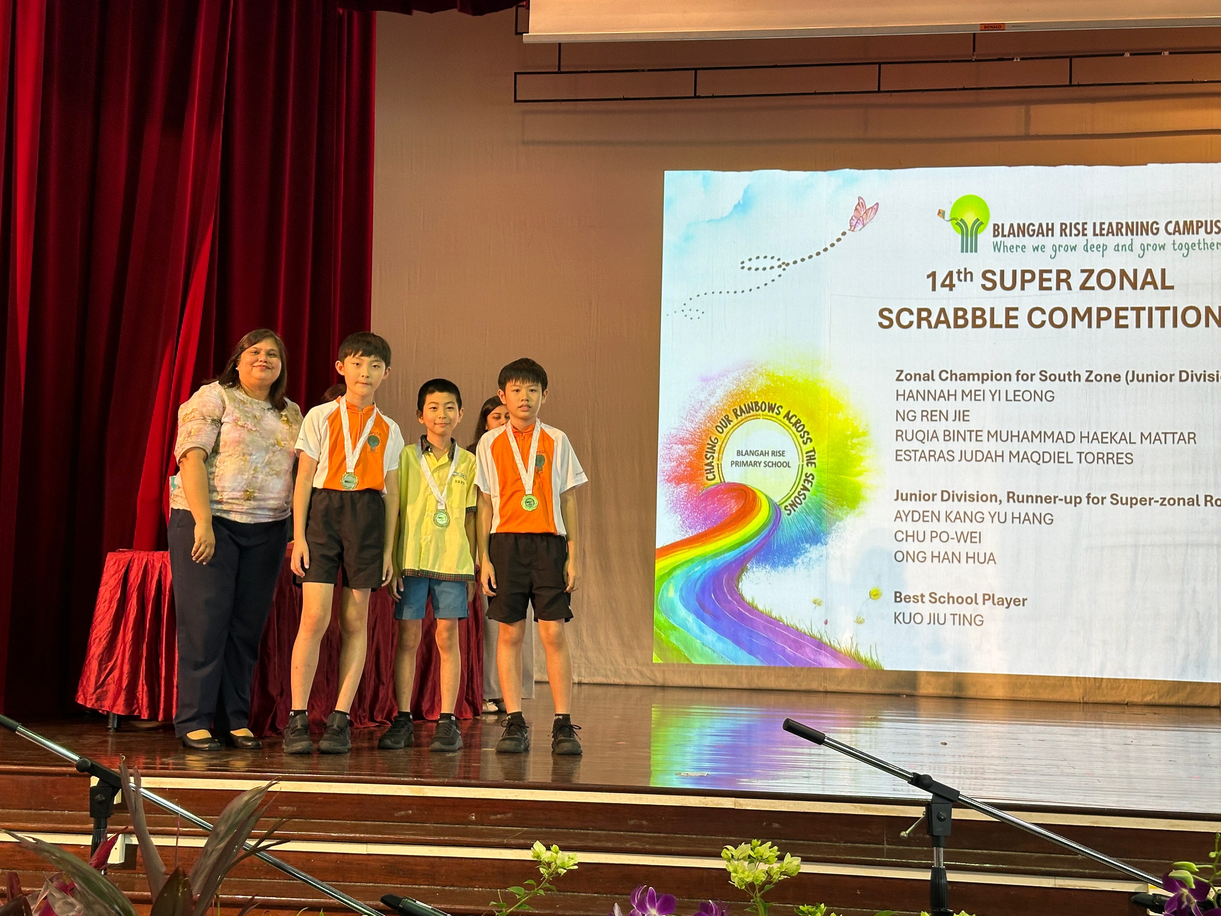 Three boys wearing medals stand on stage with an adult woman at the 14th Super Zonal Scrabble Competition.