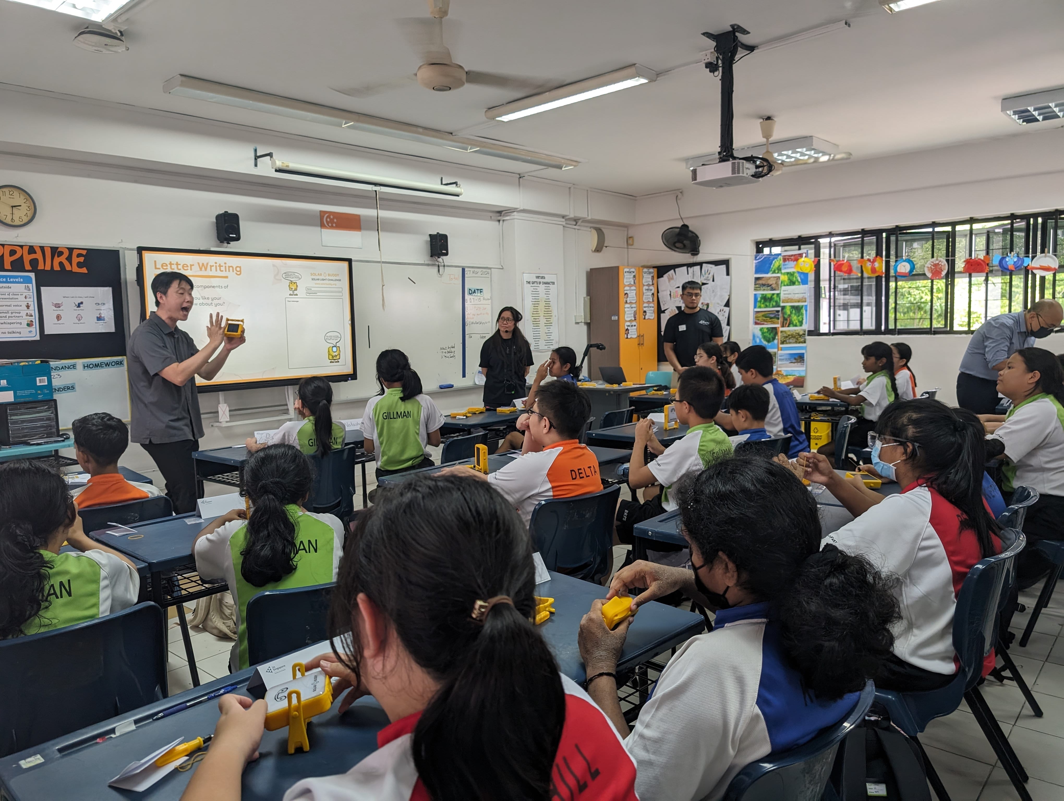 A teacher holds a yellow device while presenting a Letter Writing lesson to students seated at desks in a classroom.