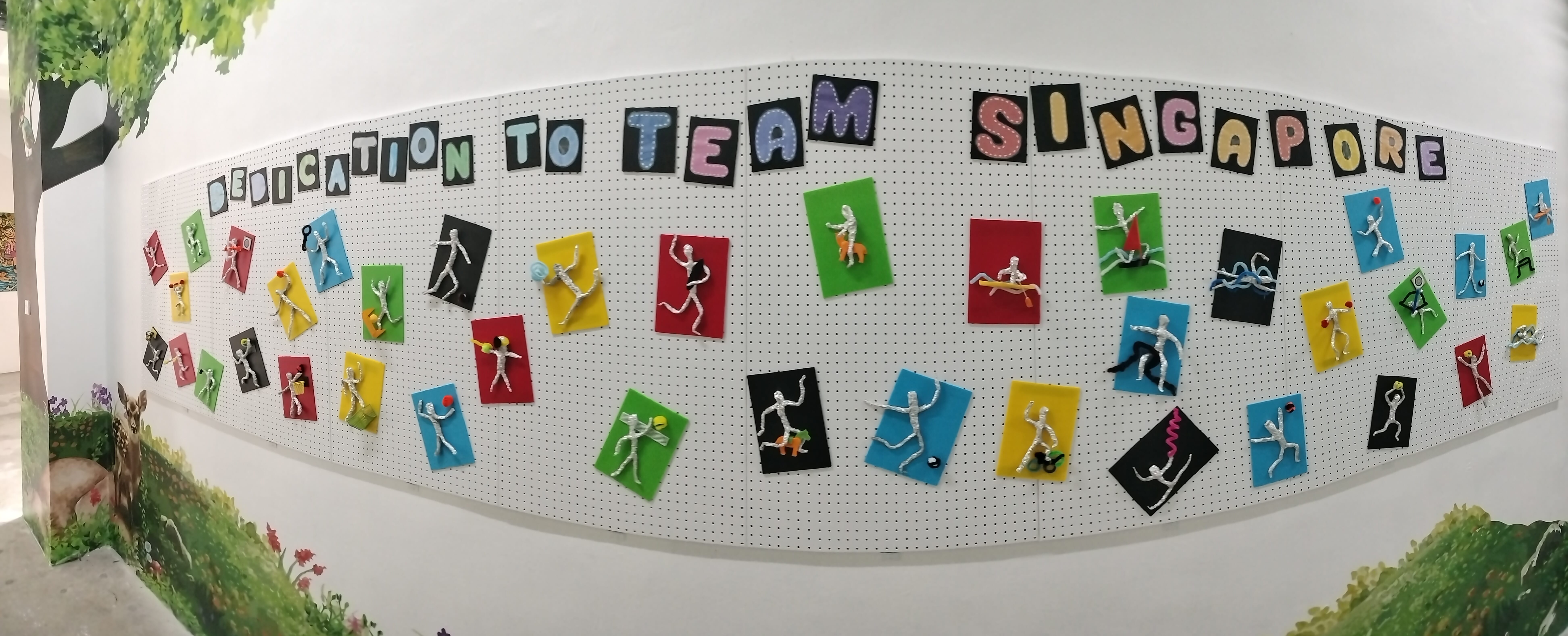 A wall display reading "Dedication to Team Singapore" with colorful cards featuring wire athlete figurines in various sports poses.