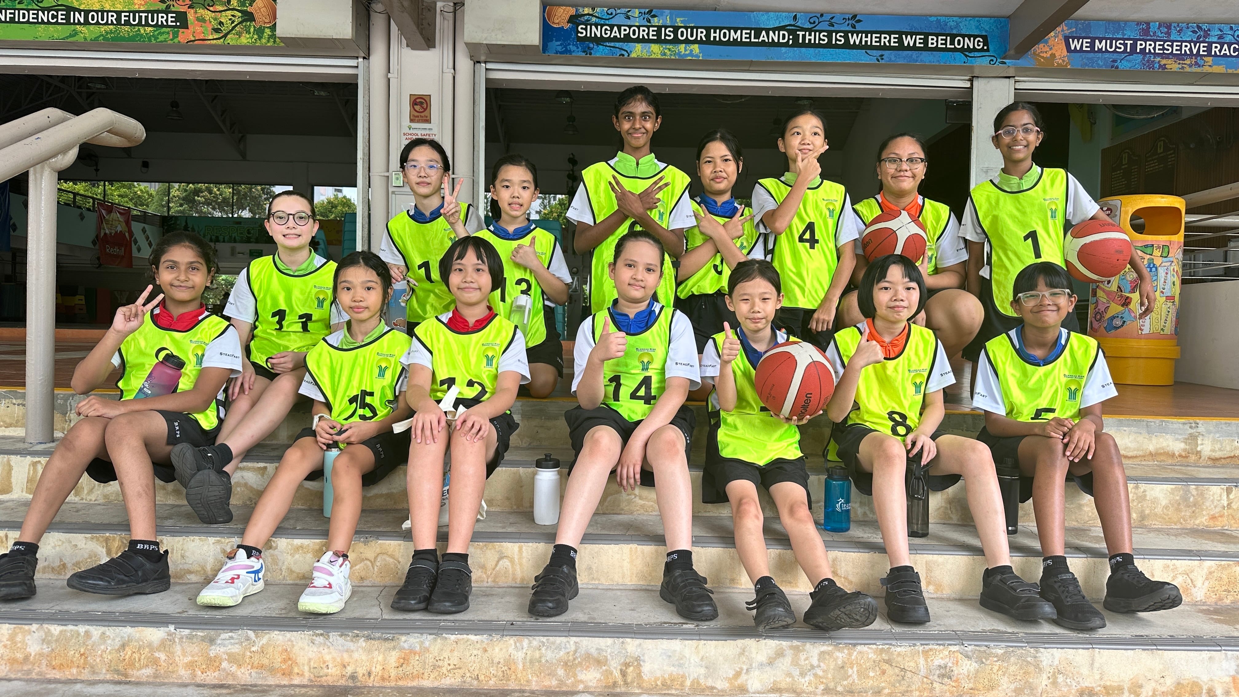 Group of young students in neon yellow numbered vests posing on school steps with basketballs in Singapore.