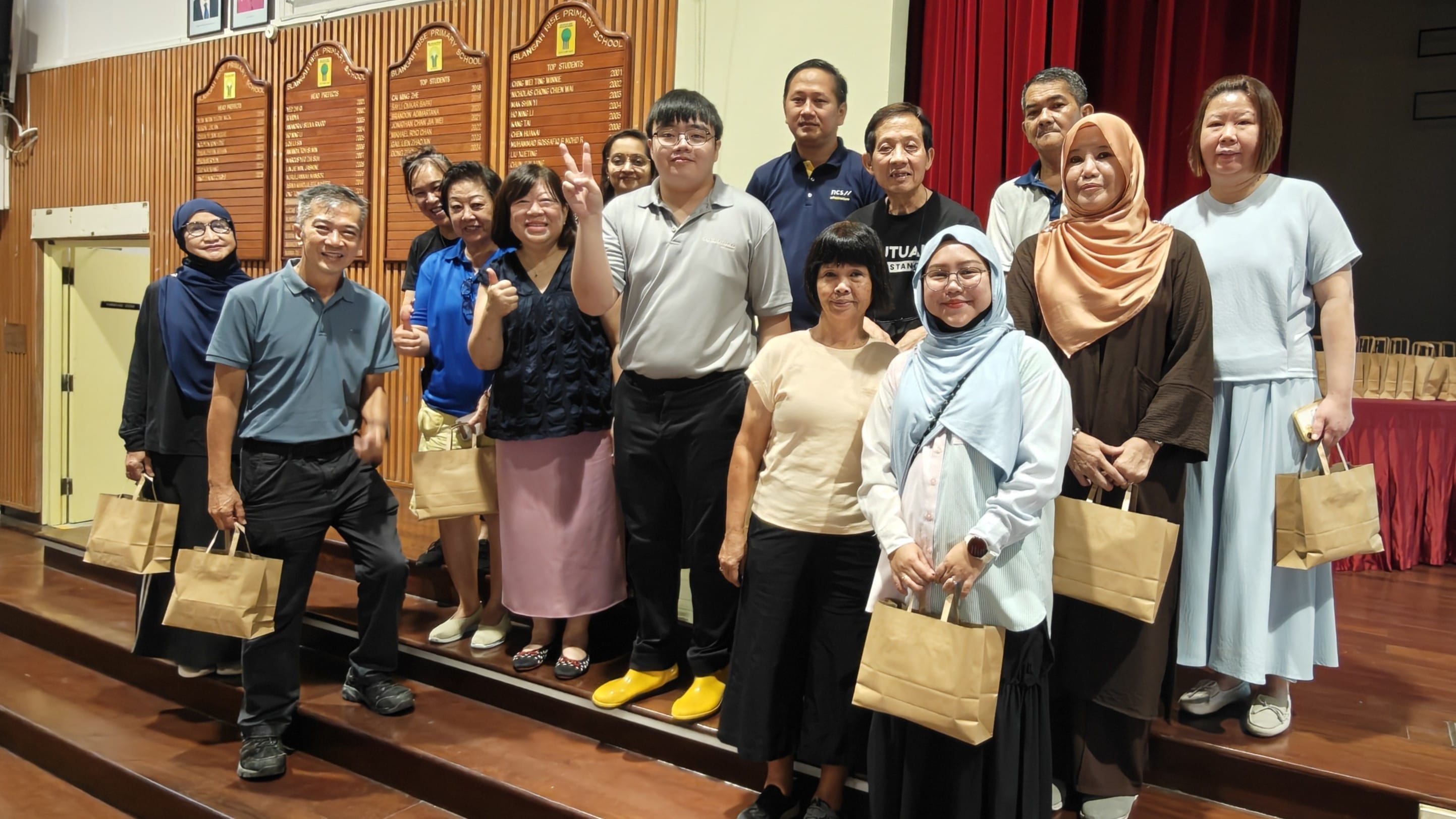 A group of about twelve adults posing together in a school hall, several holding brown paper bags.