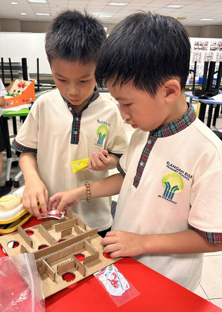 Two boys in Blangah Rise Primary School uniforms build a cardboard maze on a red table.