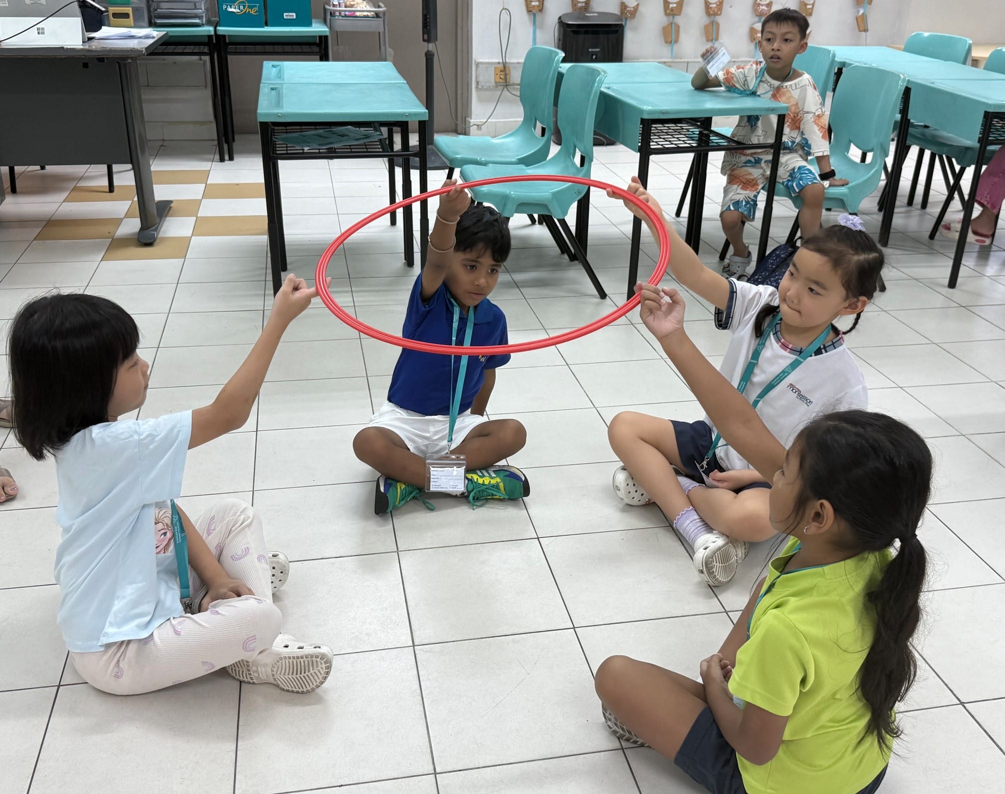 Four children sit on a classroom floor holding a red hoop together, with teal chairs and desks in the background.