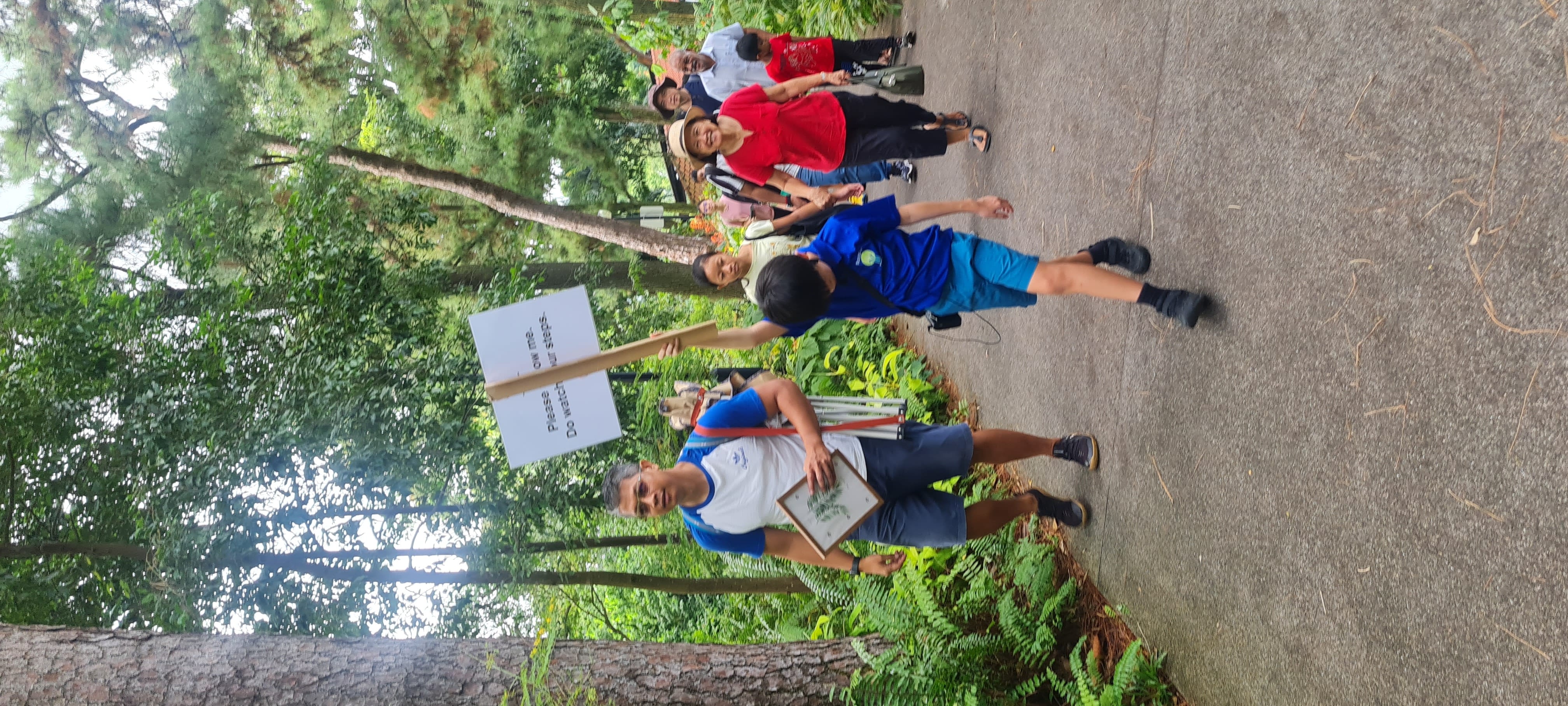 A child in blue holds a sign reading "Please follow me. Do watch your steps." leading a group on a park path.