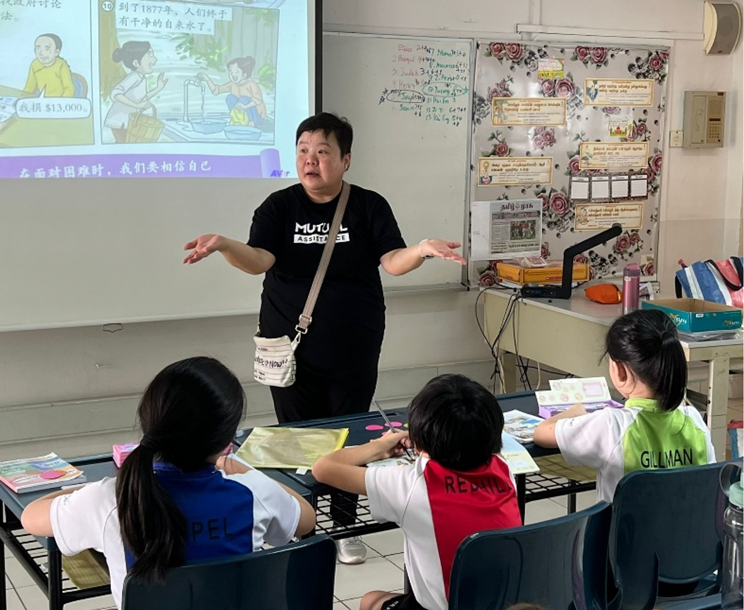 A teacher gestures while presenting a Chinese-language slideshow to young students seated at desks in a classroom.