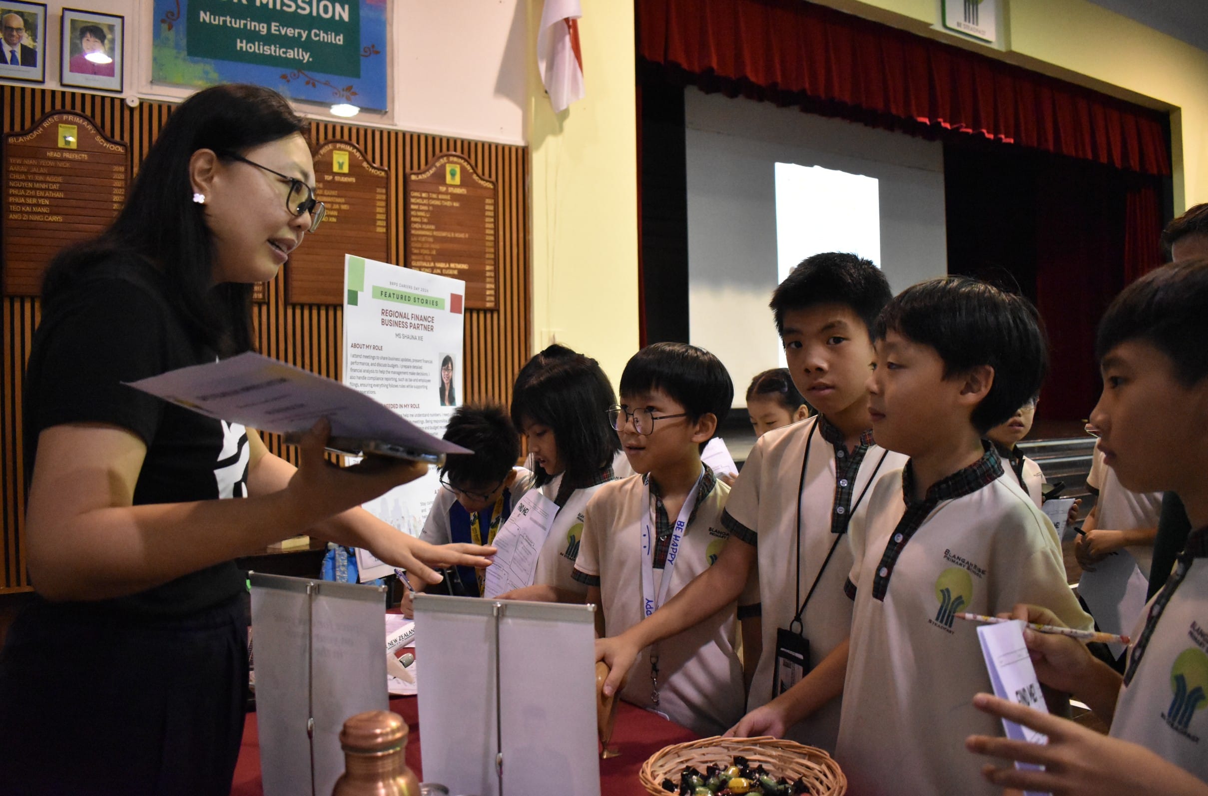 A woman in glasses speaks to a group of uniformed primary school students at an indoor school career fair booth.