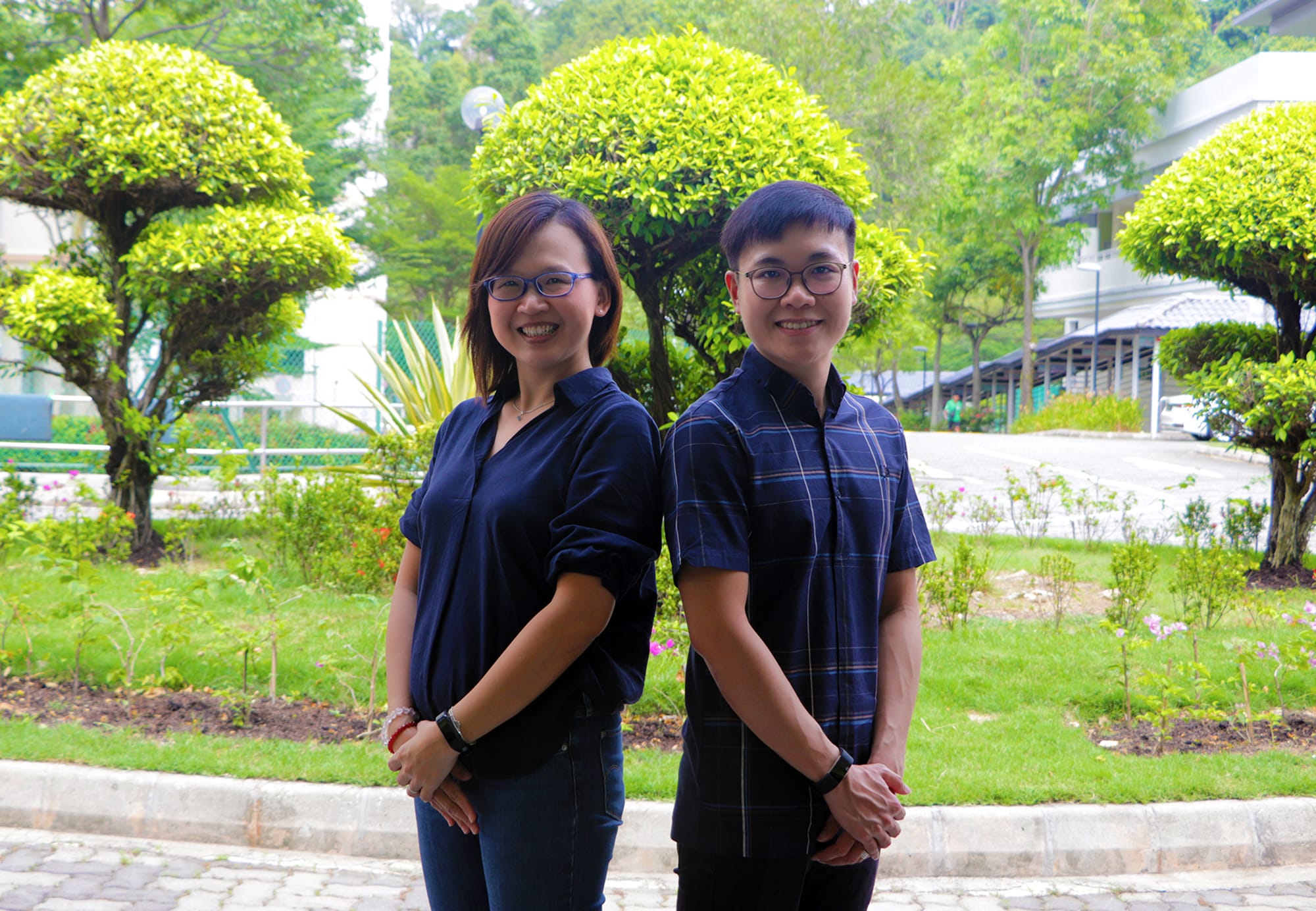 Two smiling people wearing glasses and dark blue shirts stand outdoors in a green park with trimmed round trees.