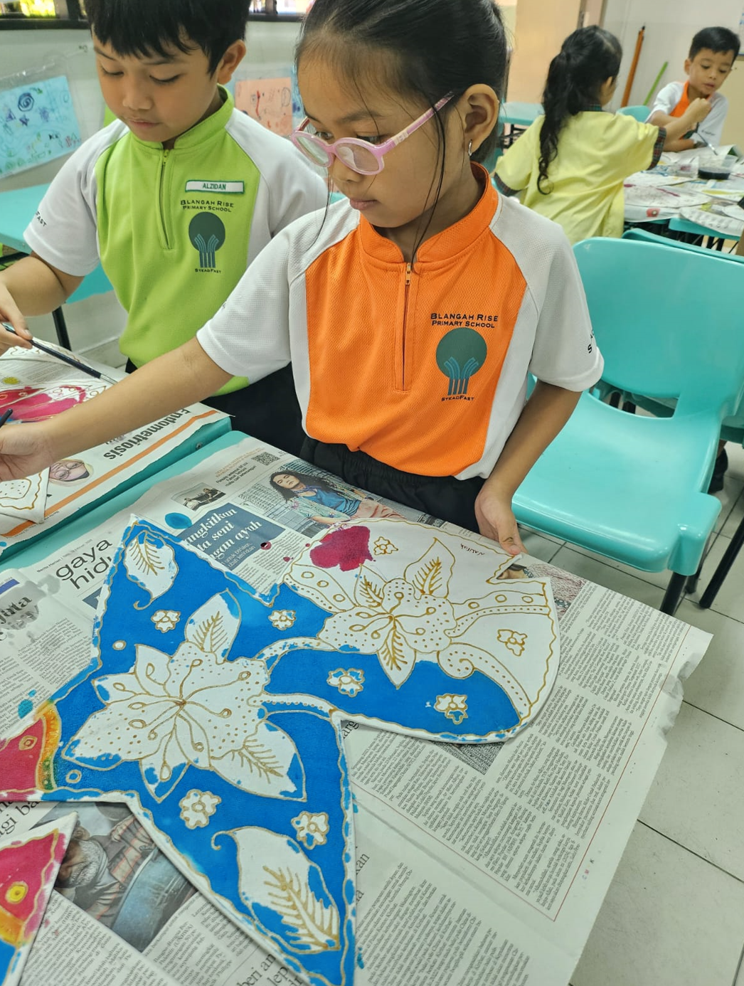 Two Blangah Rise Primary School students work on blue and white batik fabric art at a newspaper-covered table.