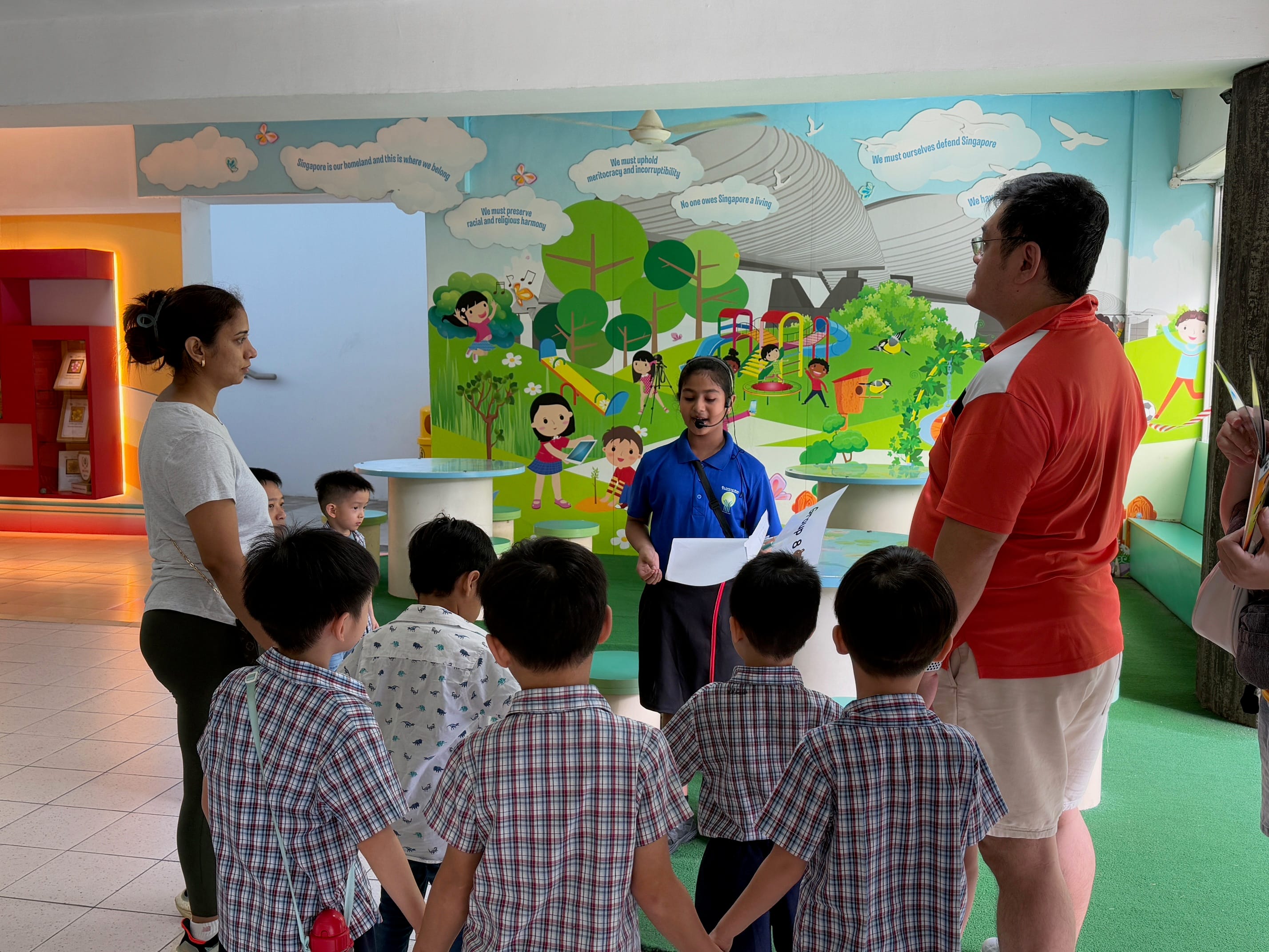 A girl in a blue uniform speaks to a group of young children in front of a colorful Singapore-themed mural.
