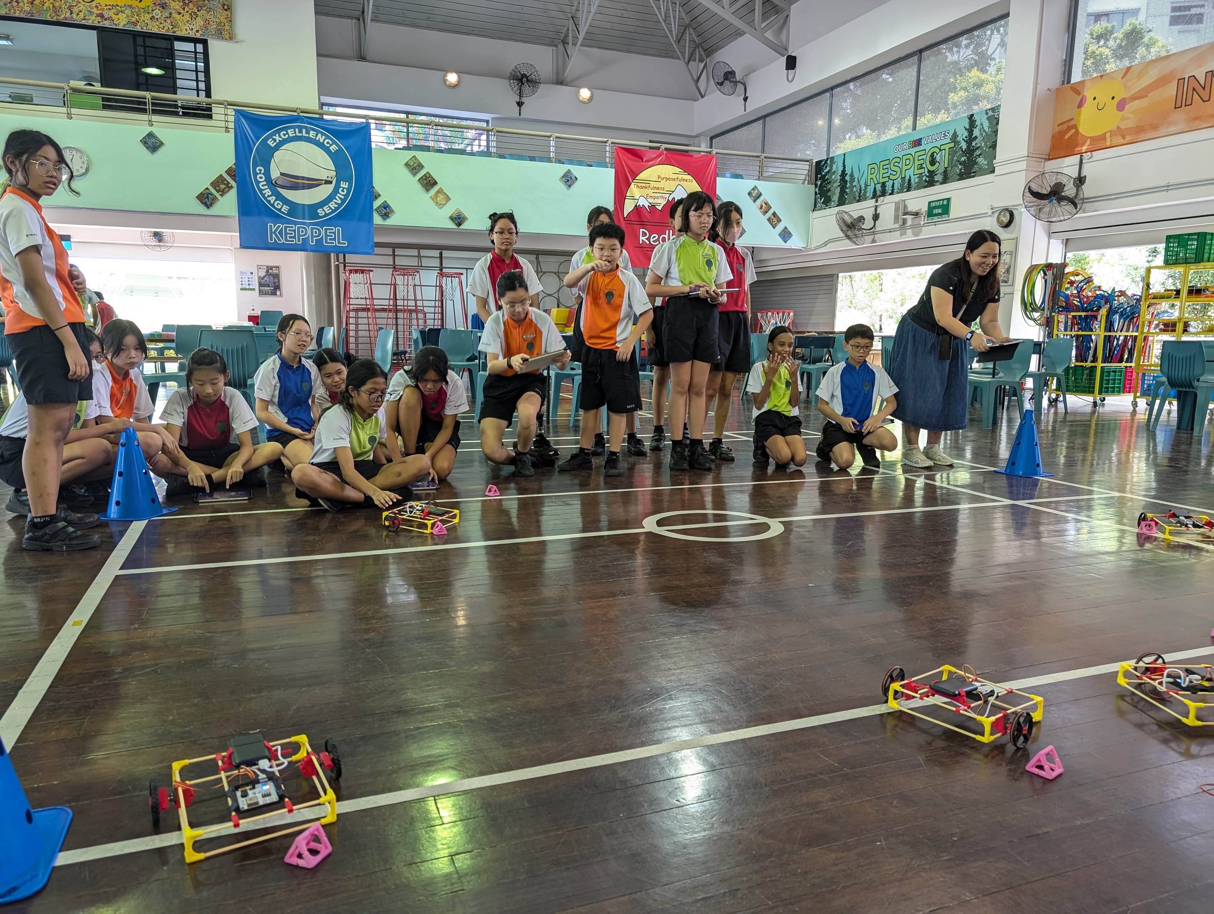 Students and a teacher watch small robotic cars on a school hall floor during a robotics competition event.