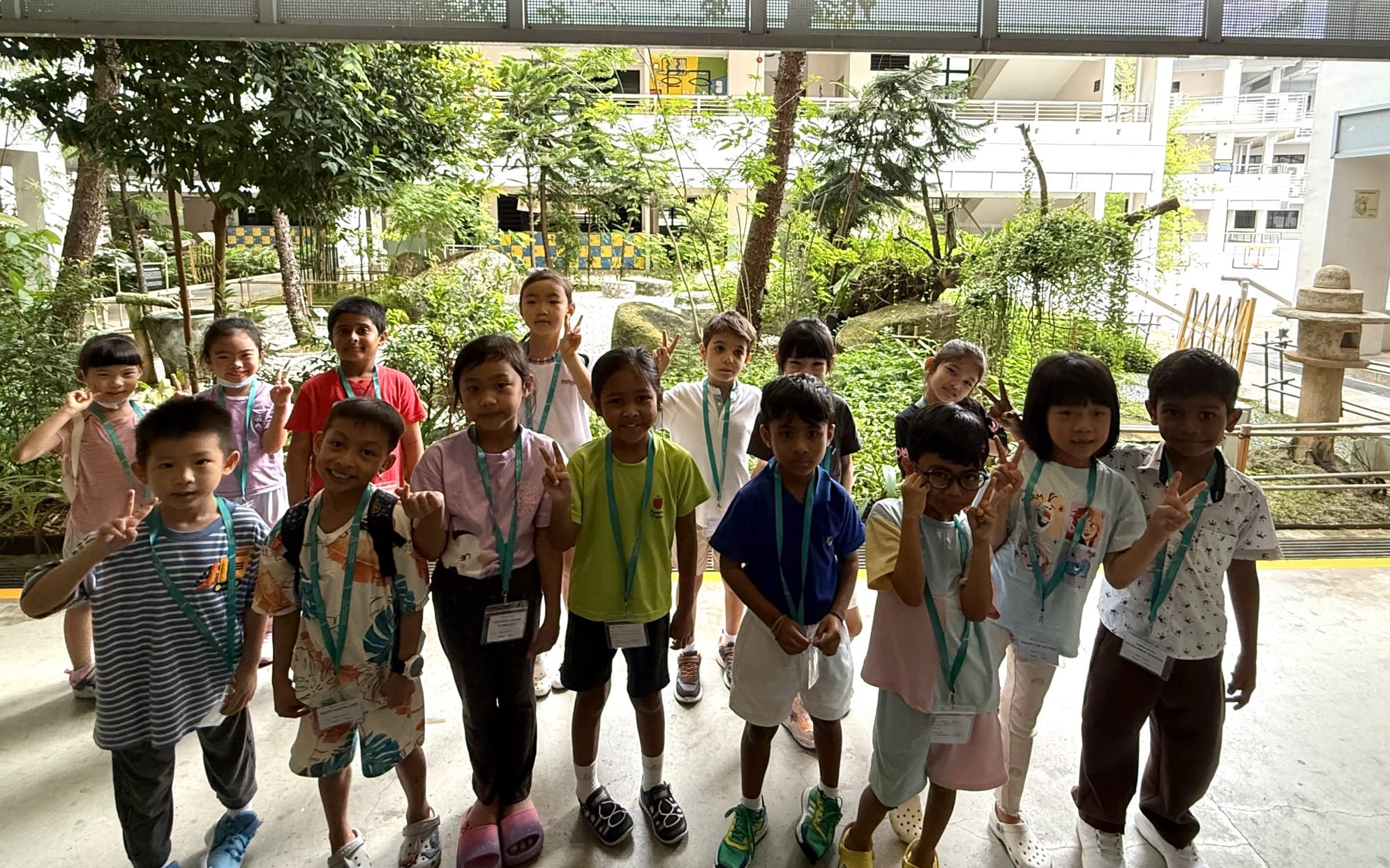 A group of young children wearing lanyards pose with peace signs in a school courtyard surrounded by trees.