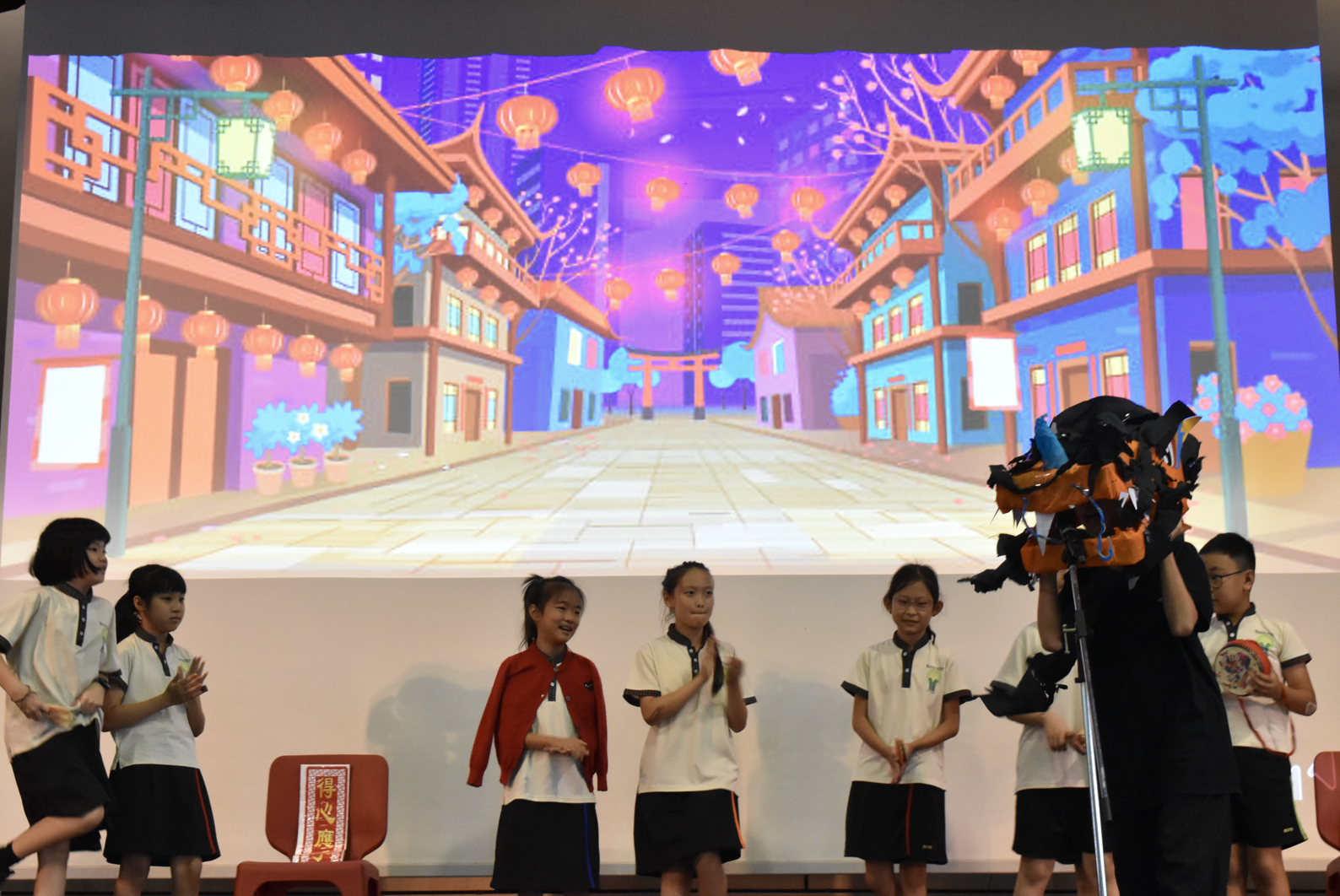Children in school uniforms perform on stage before a projected backdrop of a colorful Chinese street with red lanterns.