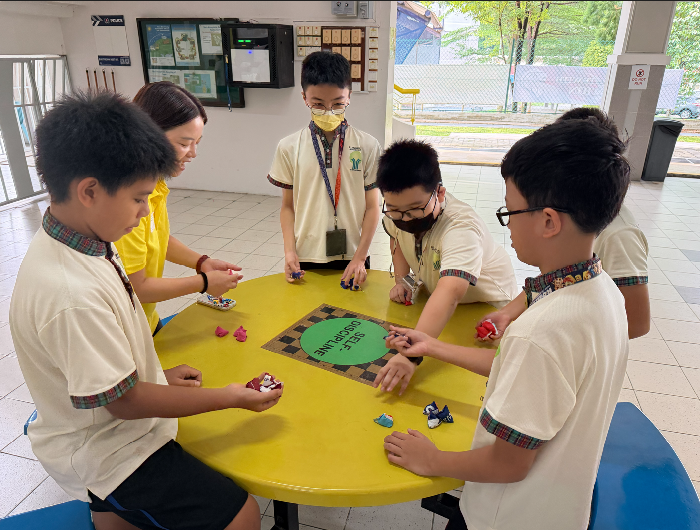 Five school children in uniforms gather around a yellow round table with a self-discipline board game.