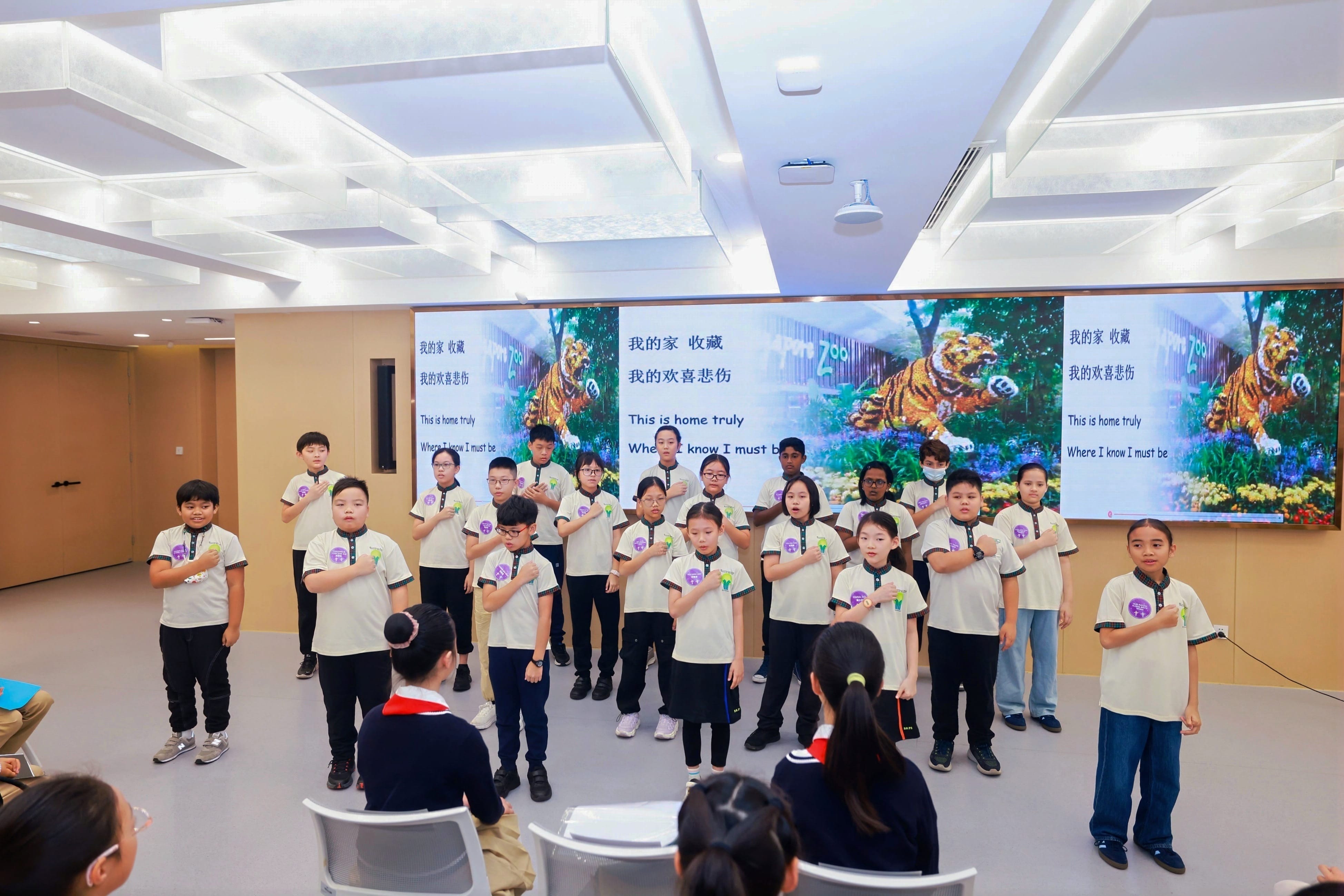 A group of children in matching white shirts perform a choir in front of a screen displaying Chinese and English lyrics.