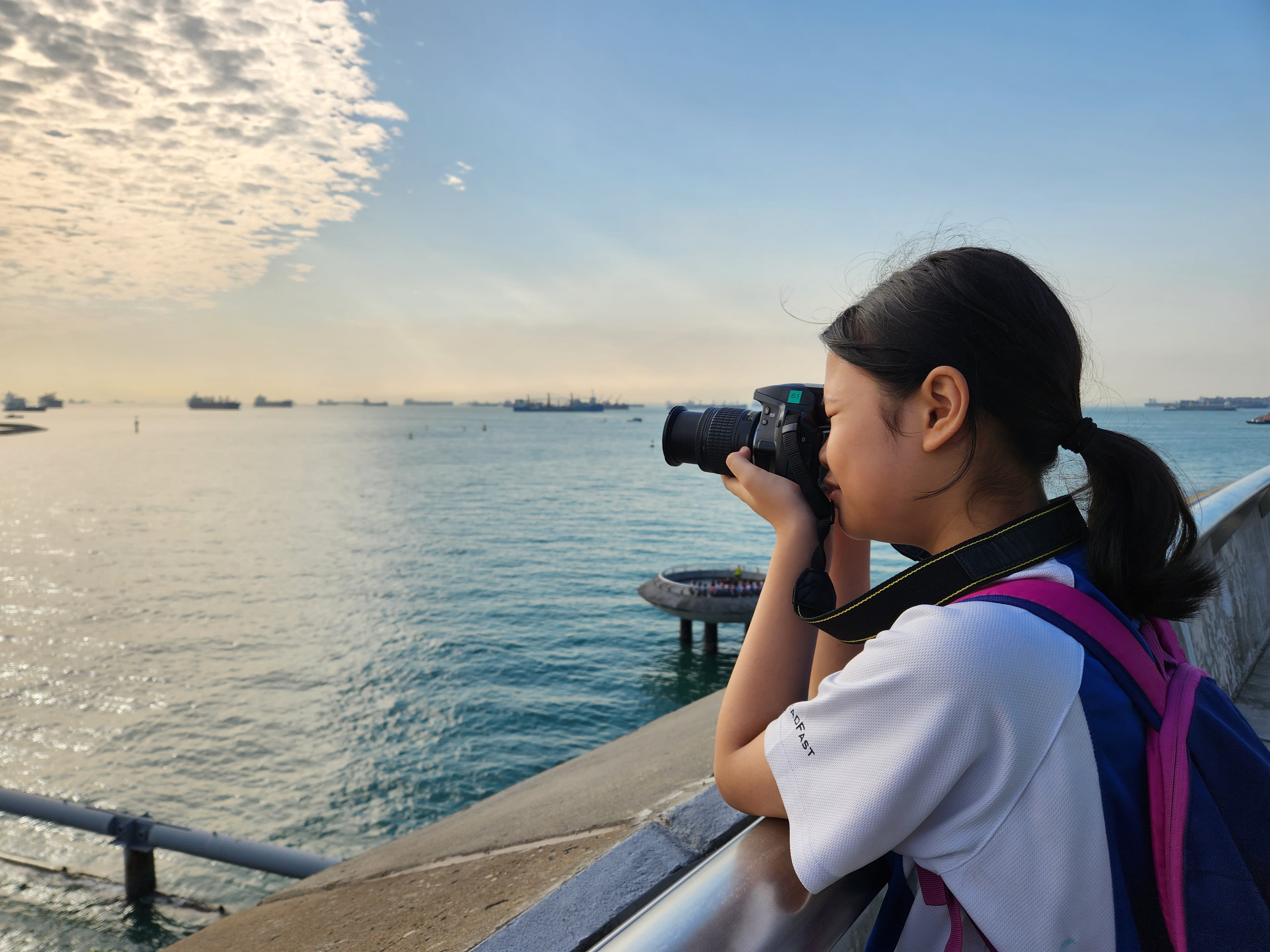 Young girl with a ponytail and backpack photographing a calm sea with ships under a partly cloudy sky.