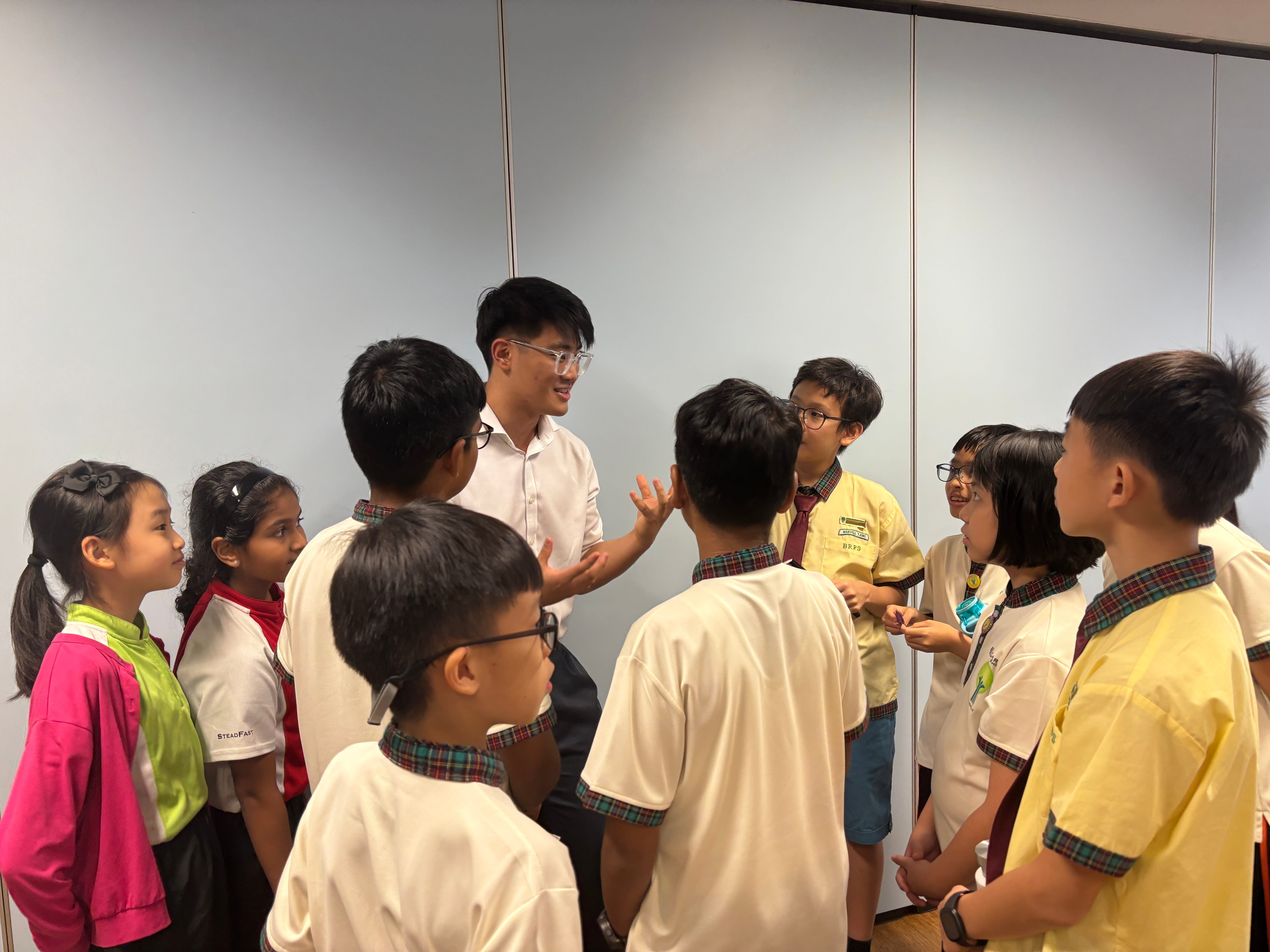 Teacher in white shirt talks to a group of schoolchildren in uniform gathered in a circle indoors.