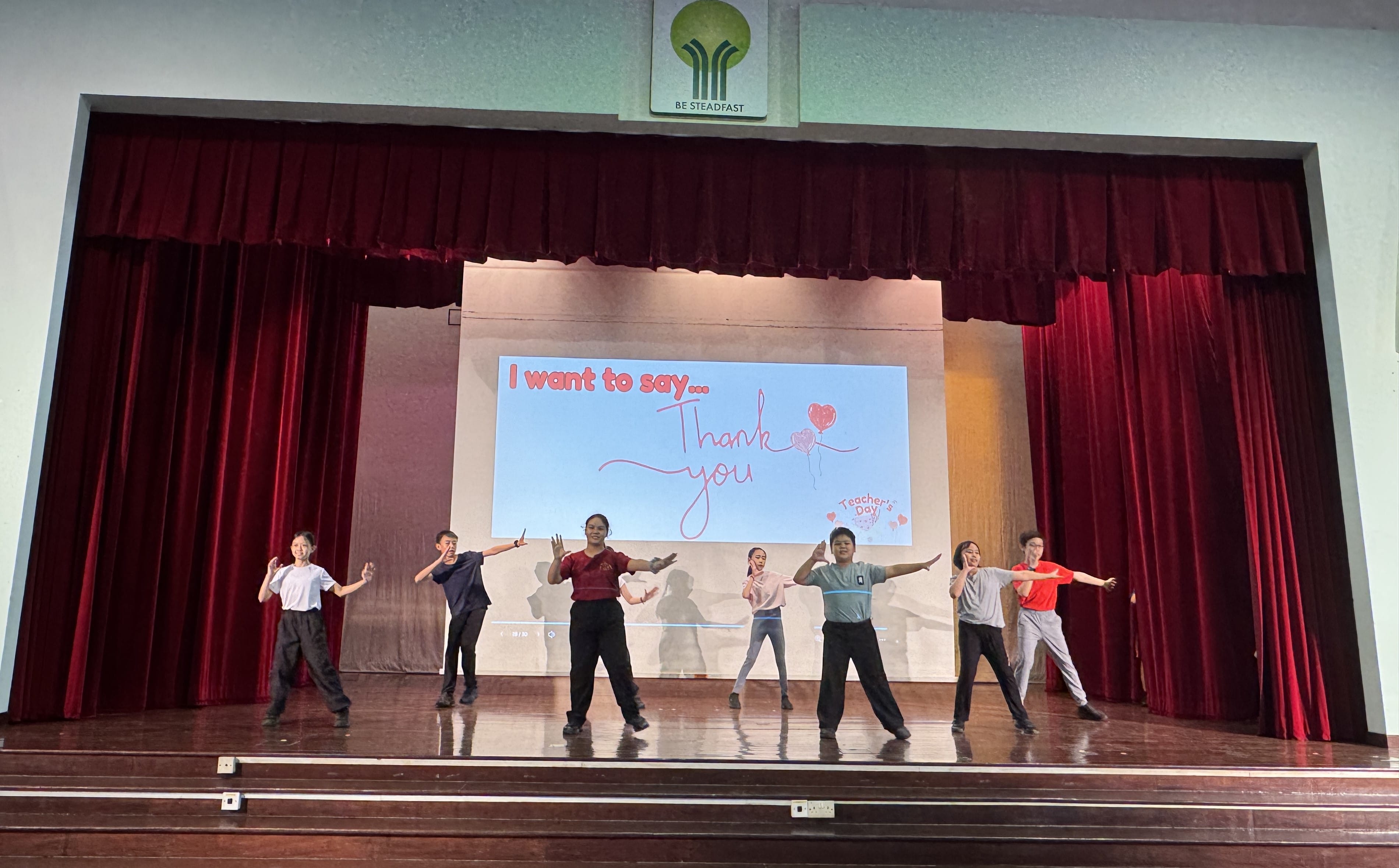 Seven people dance on a stage with red curtains; screen behind reads I want to say... Thank you.