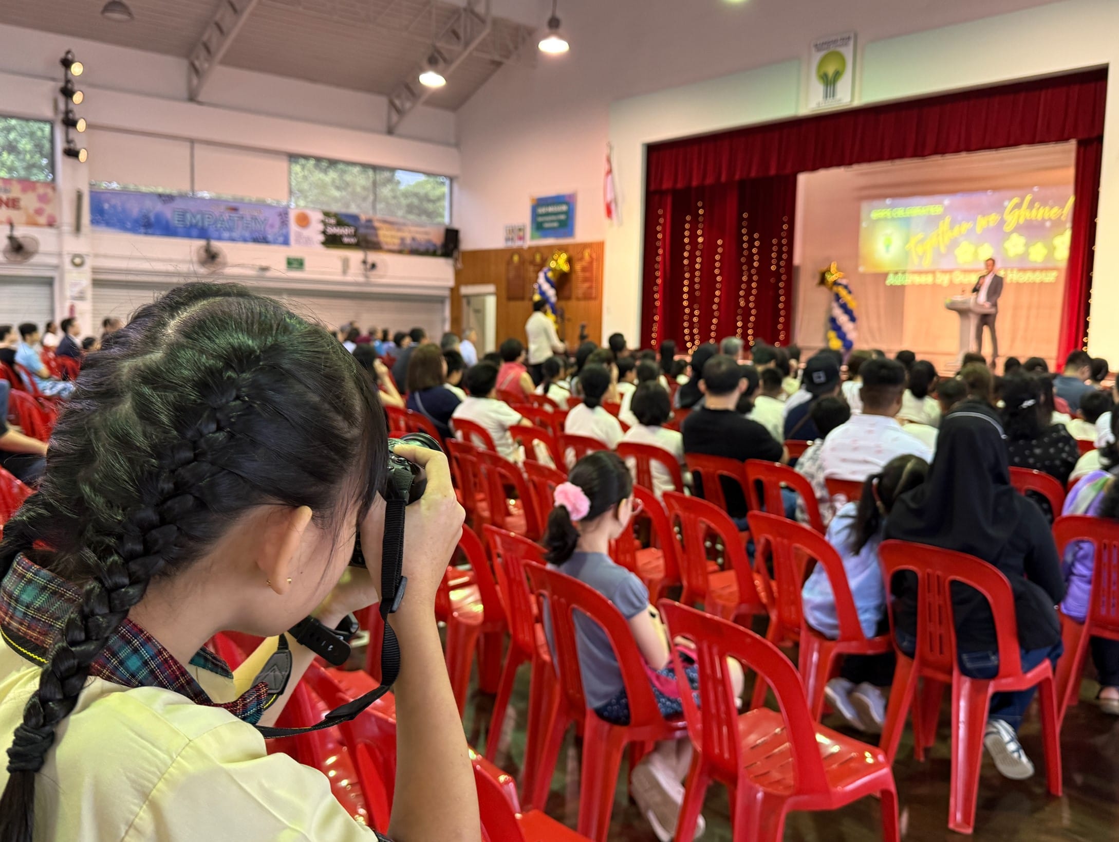 A girl with braided hair photographs a school hall event with red chairs and a speaker on stage with red curtains.