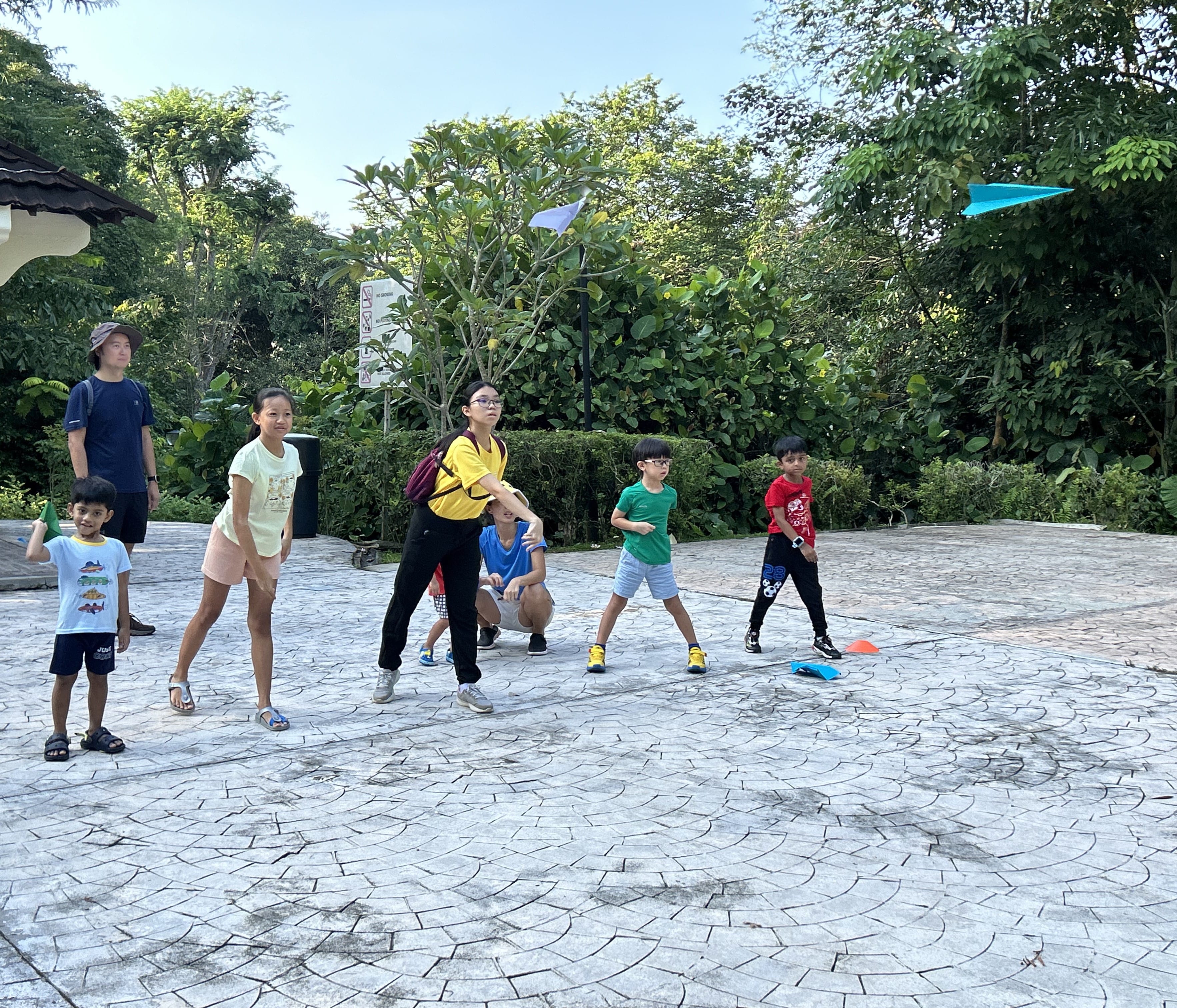 Several children and adults play with paper planes on a paved outdoor area surrounded by green trees.