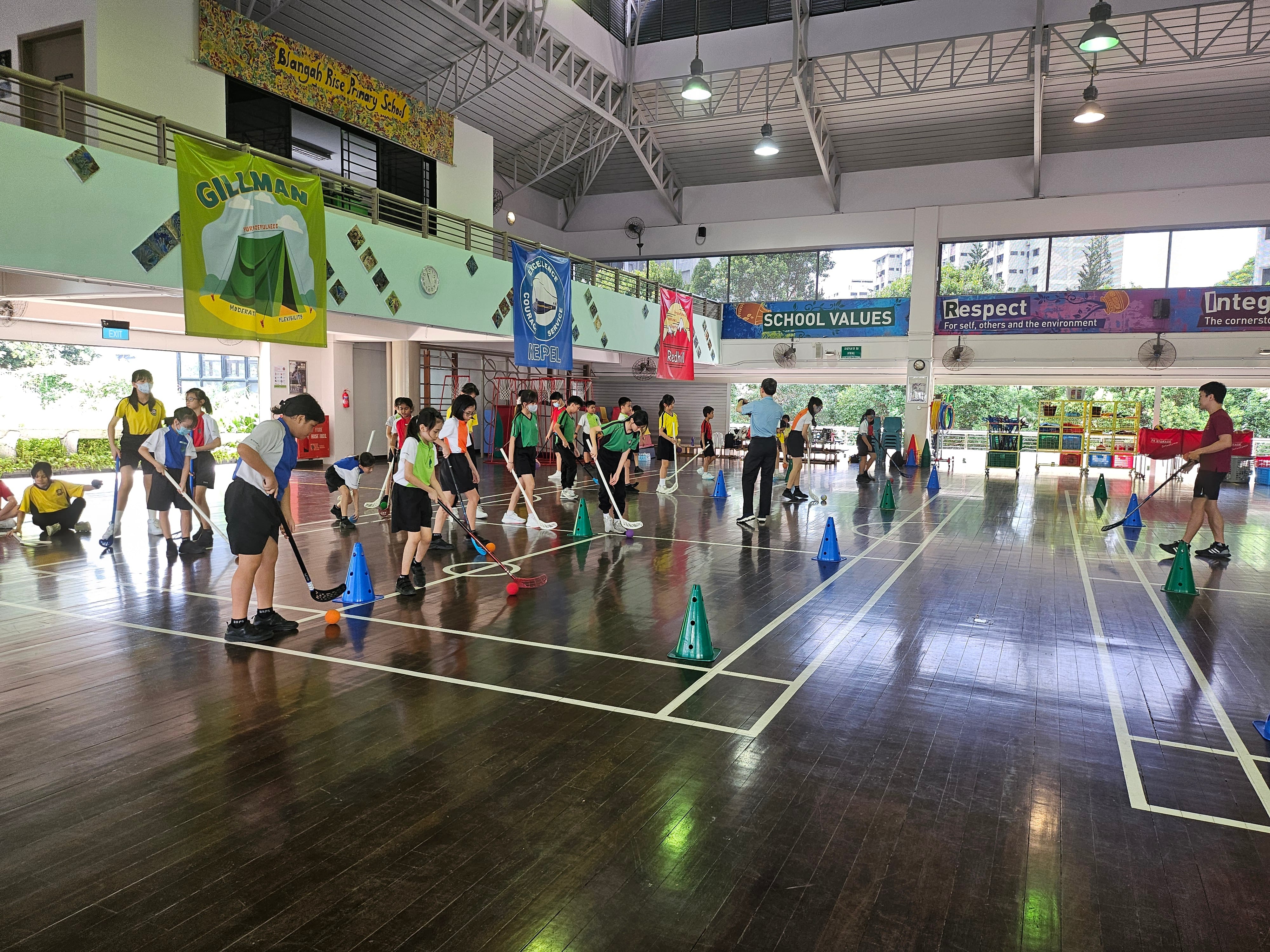 Students in a school gym practice floor hockey with sticks and balls between colored cones.