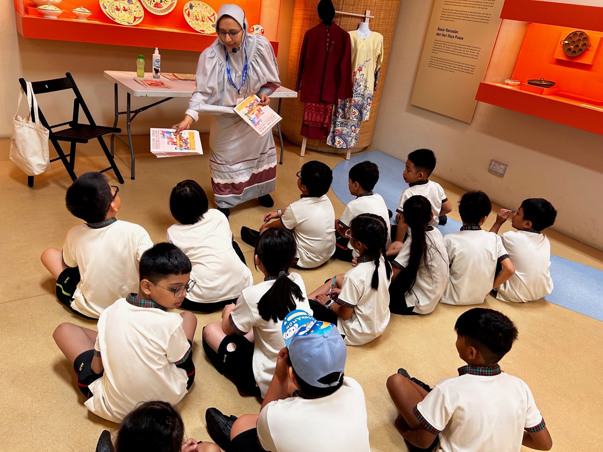 A woman in traditional dress holds books while teaching a group of young students seated on a museum floor.
