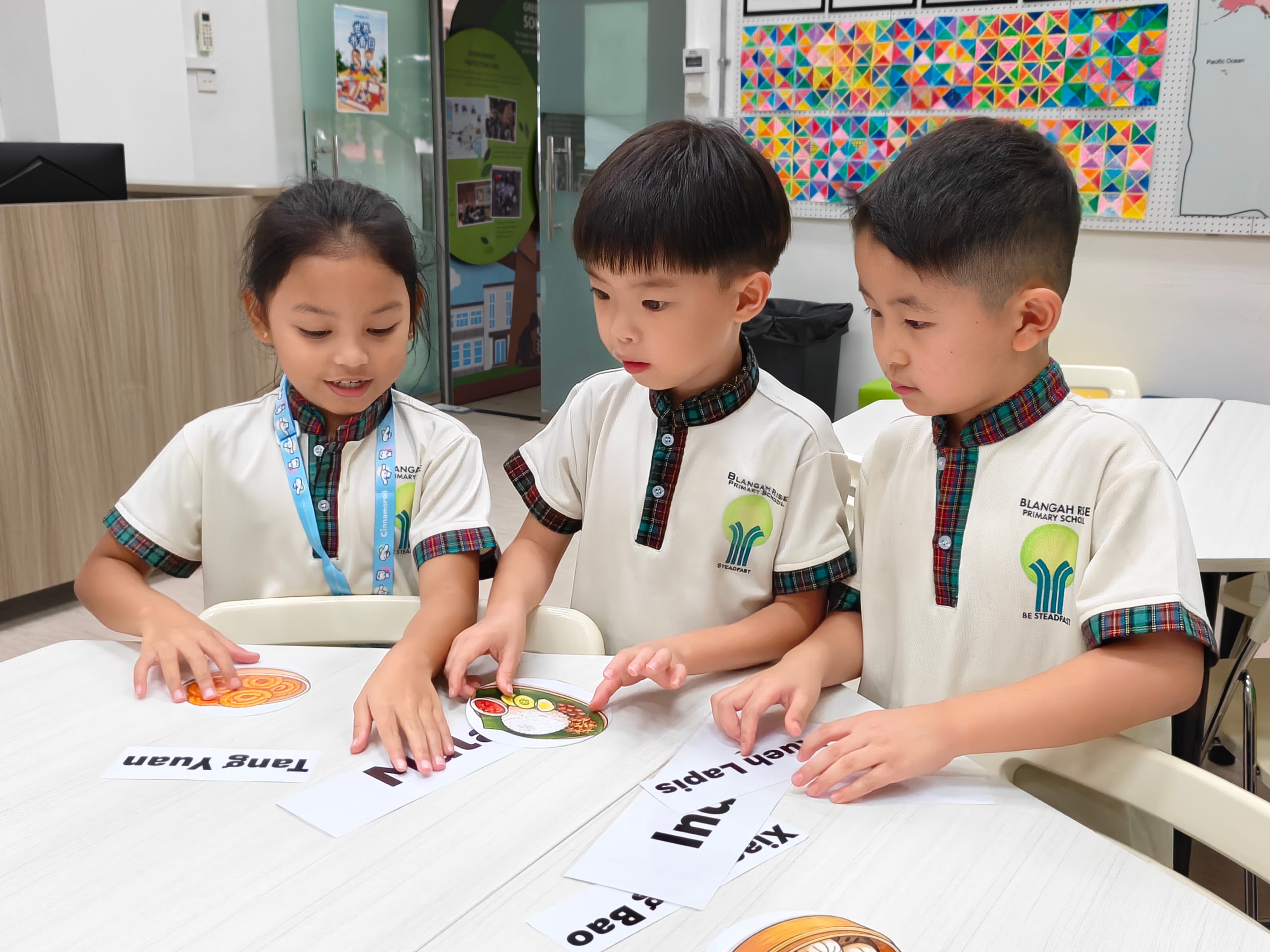 Three young students in school uniforms examine food picture cards labeled with names like Tang Yuan at a classroom table.