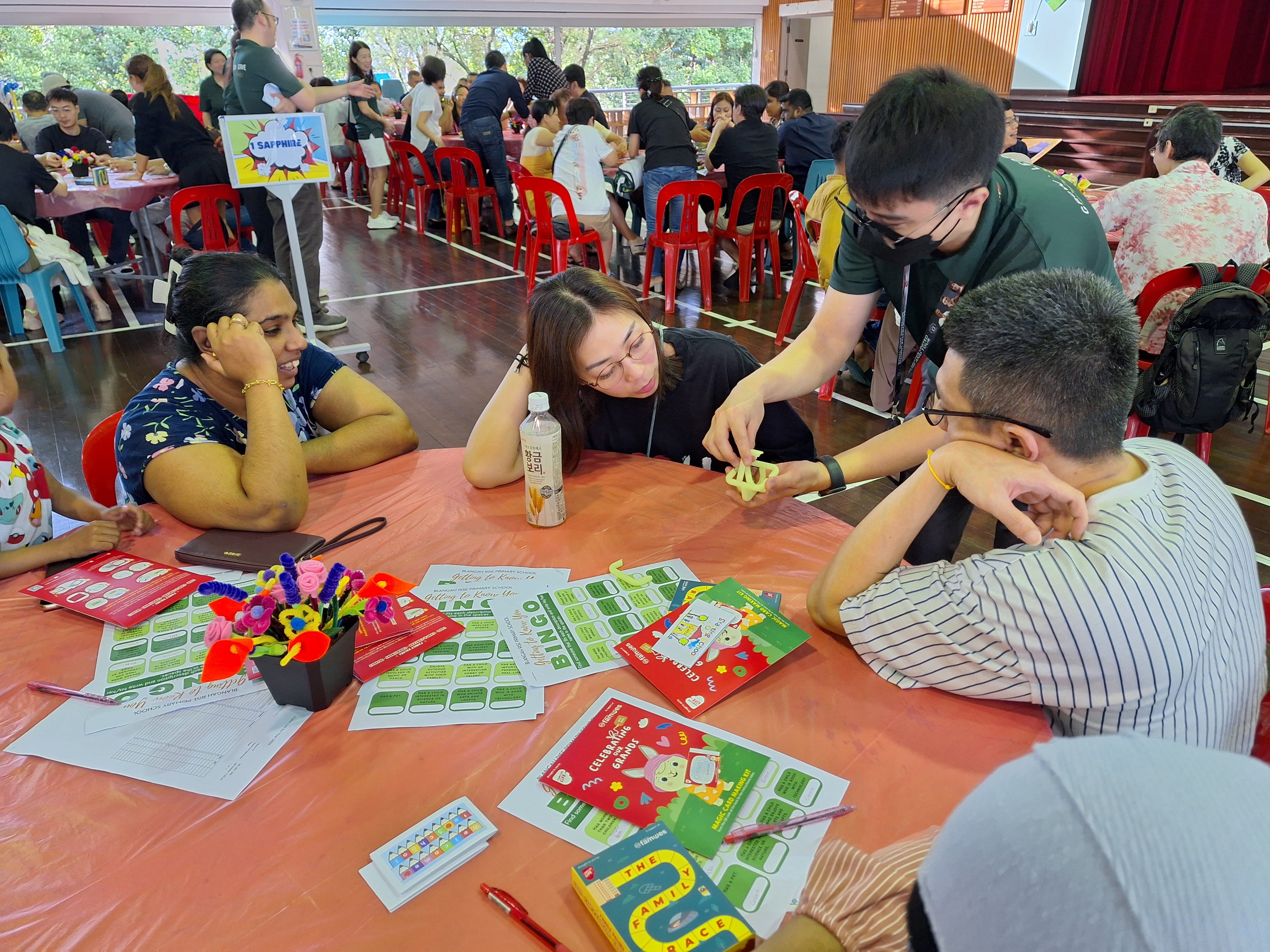 People sit around a table at a busy hall event as a man shows a small object to two others.