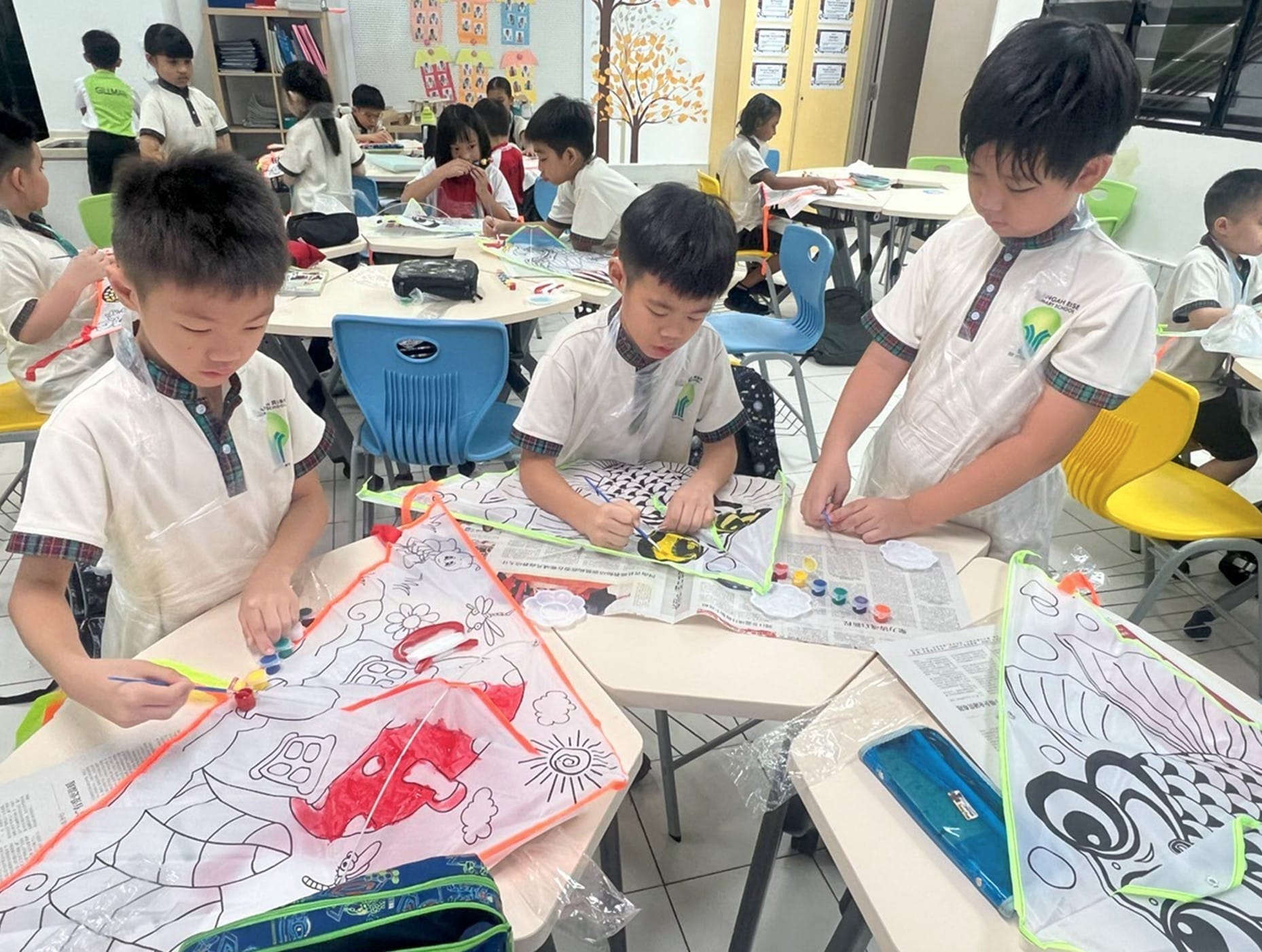 Young students in school uniforms painting kite designs with small paint pots in a bright classroom.
