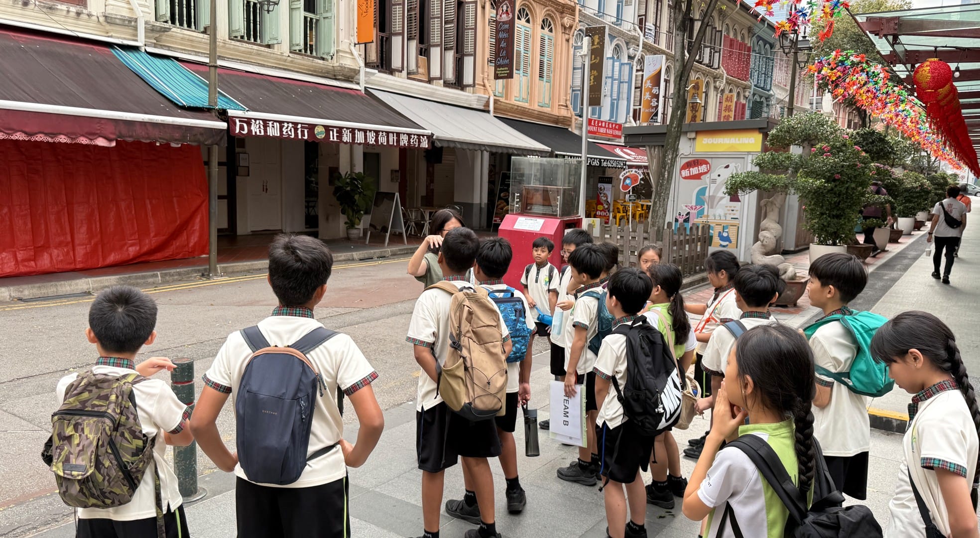 A group of school children with backpacks gathered on a street in Singapore's Chinatown with shophouses and festive decorations.