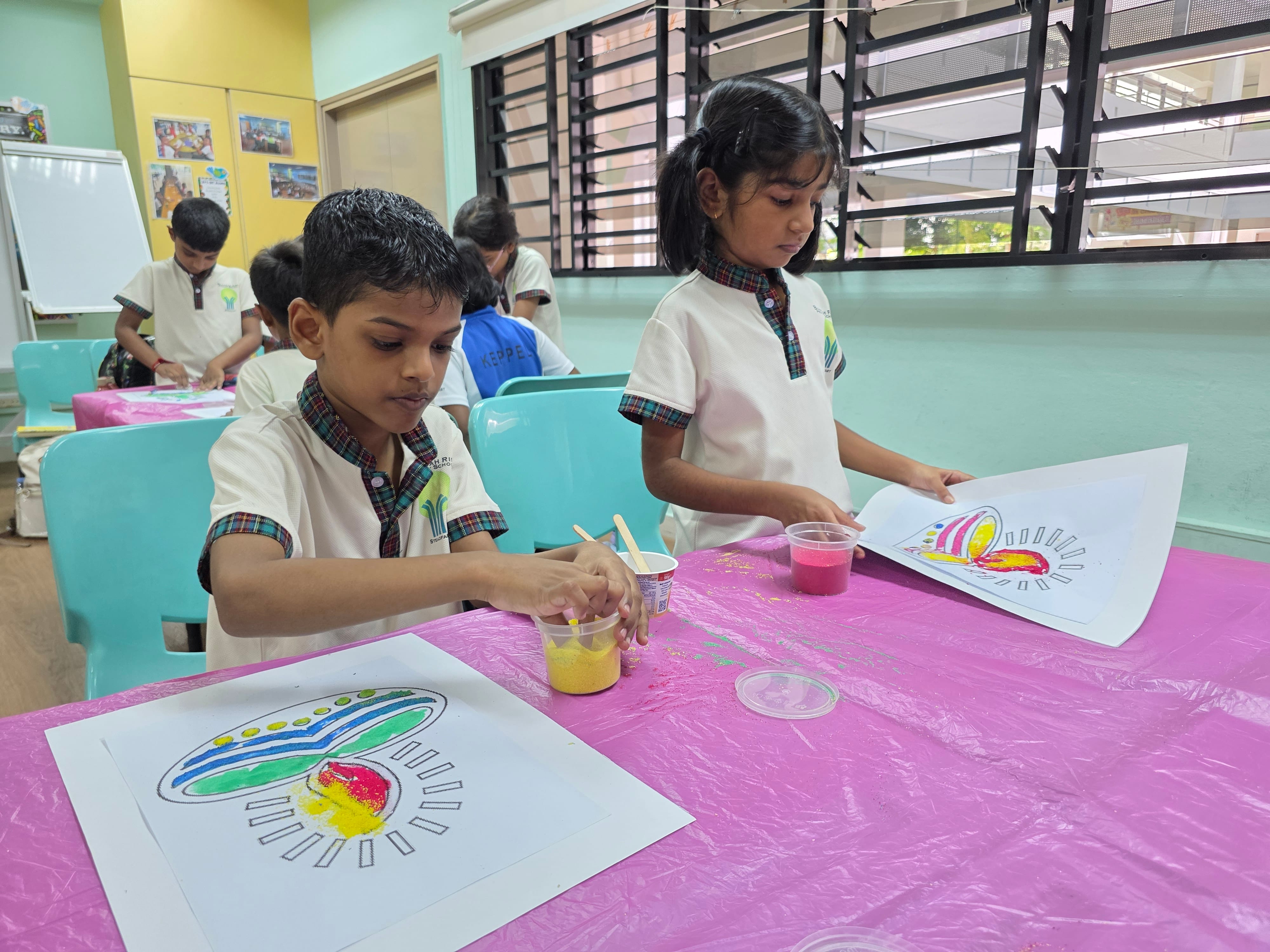 Two young students in white uniforms use colored sand to decorate drawings at a pink-covered table in a classroom.