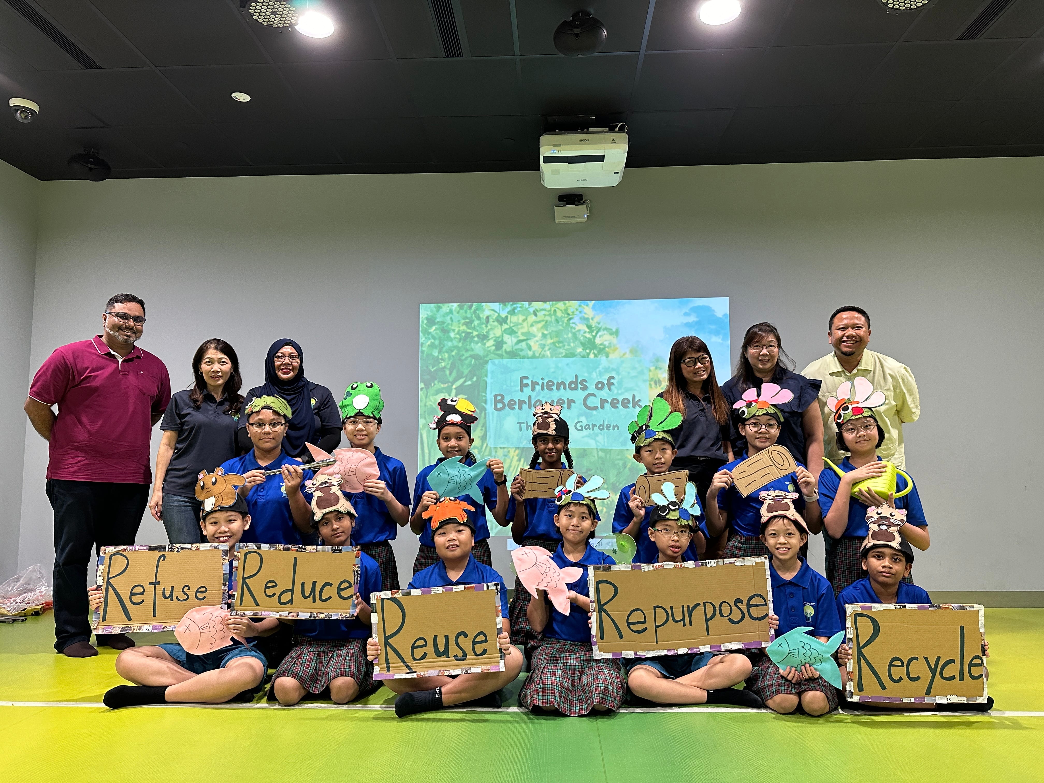 Children in blue uniforms wear animal hats and hold cardboard signs reading Refuse, Reduce, Reuse, Repurpose, Recycle with teachers.