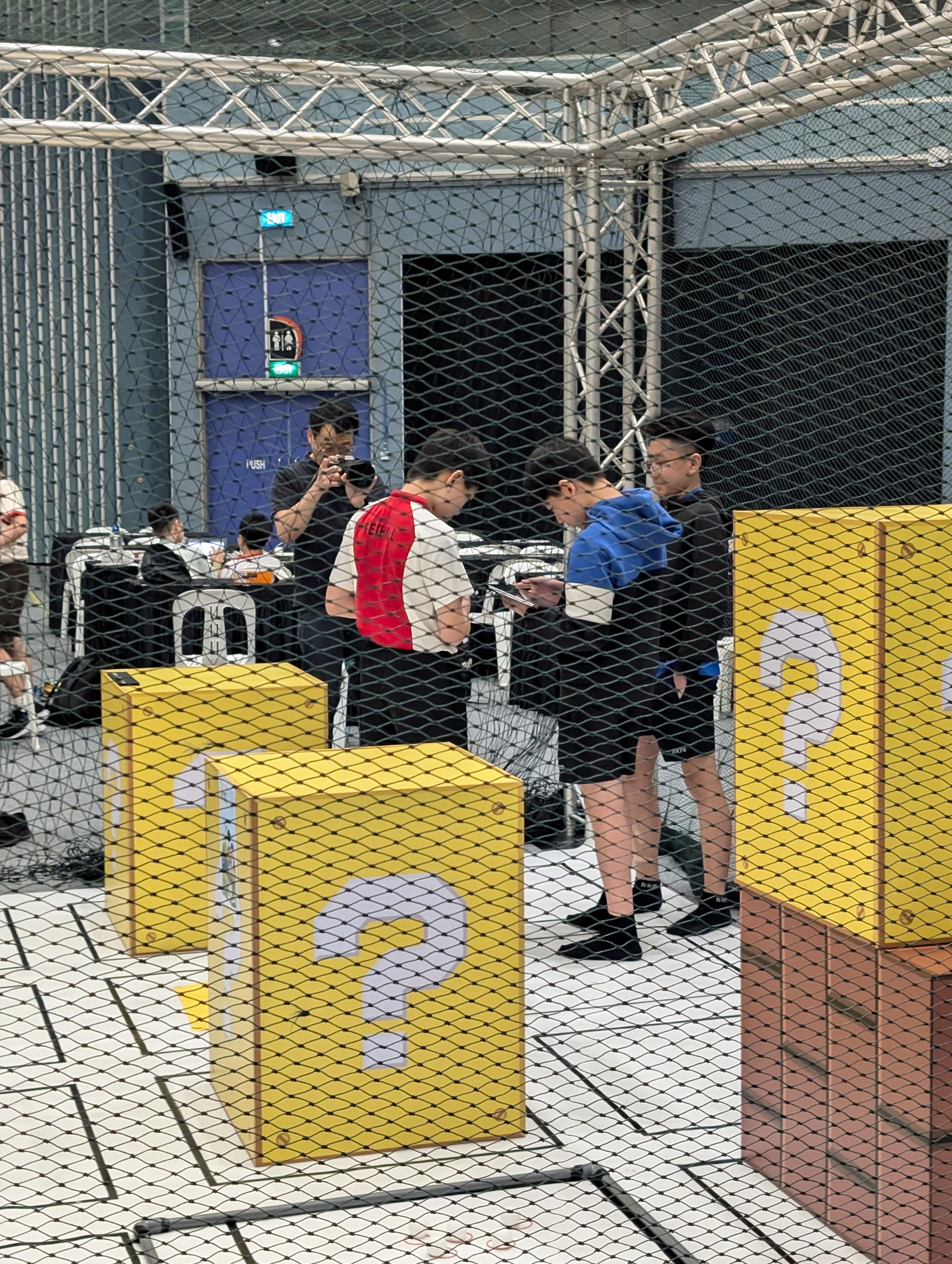Several people stand near yellow numbered boxes inside a netted indoor arena, viewed through a safety net.