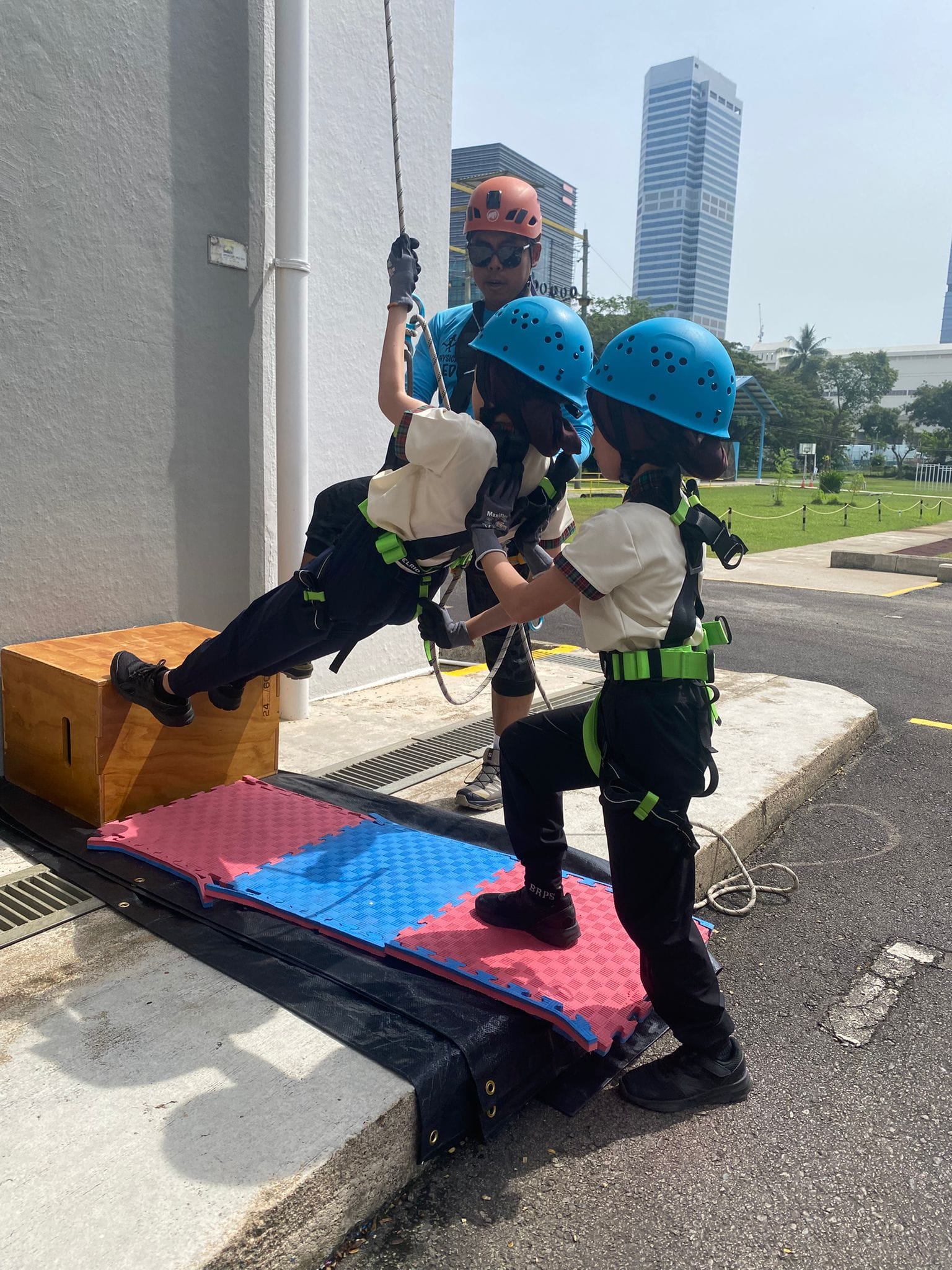 Three children in helmets and harnesses practice rappelling down a building wall outdoors near city skyscrapers.