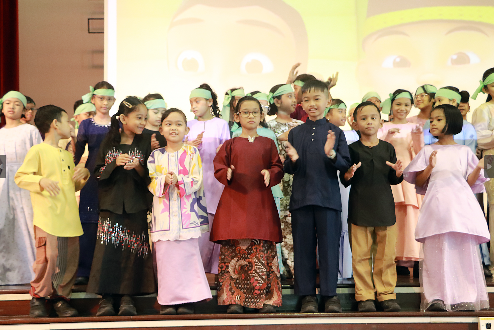 Children in colorful traditional outfits stand on stage, many wearing green headbands and clapping.