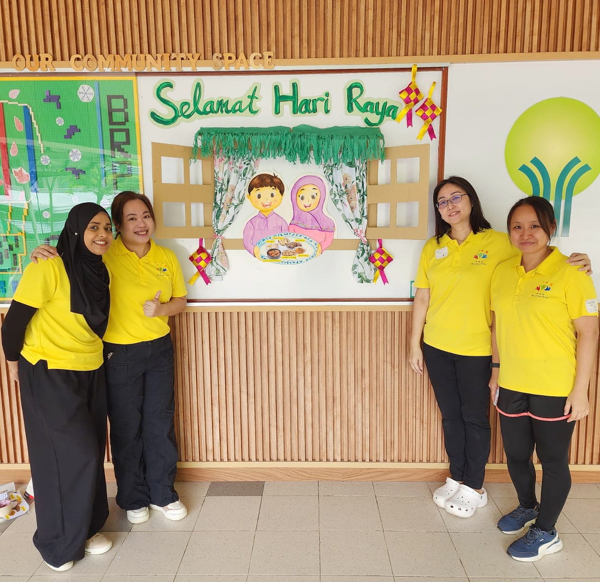 Four women in yellow shirts stand before a colorful Selamat Hari Raya display in a community space.