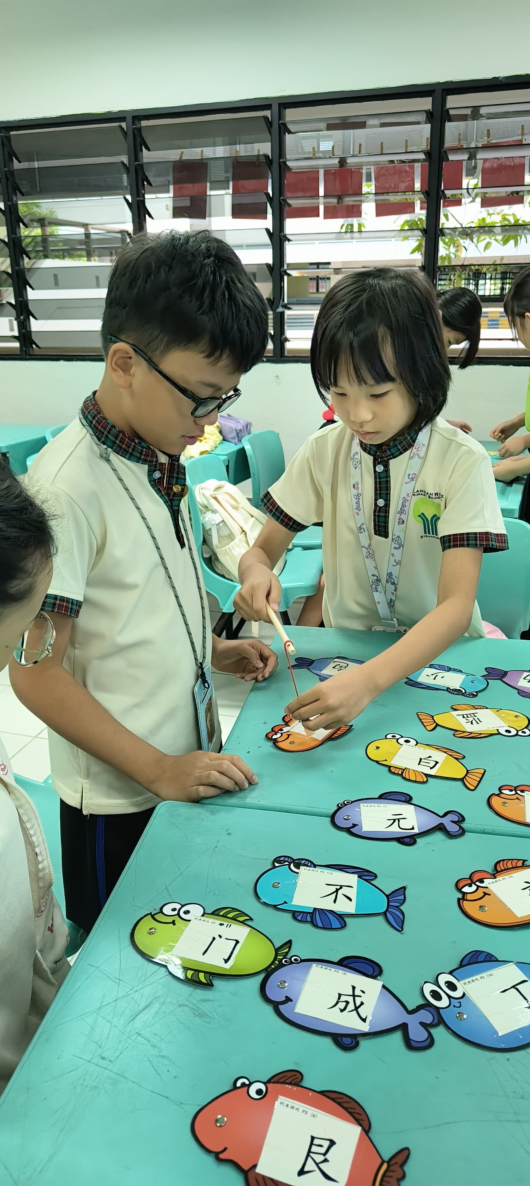 Two children in school uniforms use a magnetic wand to pick up fish-shaped cards on a classroom table.